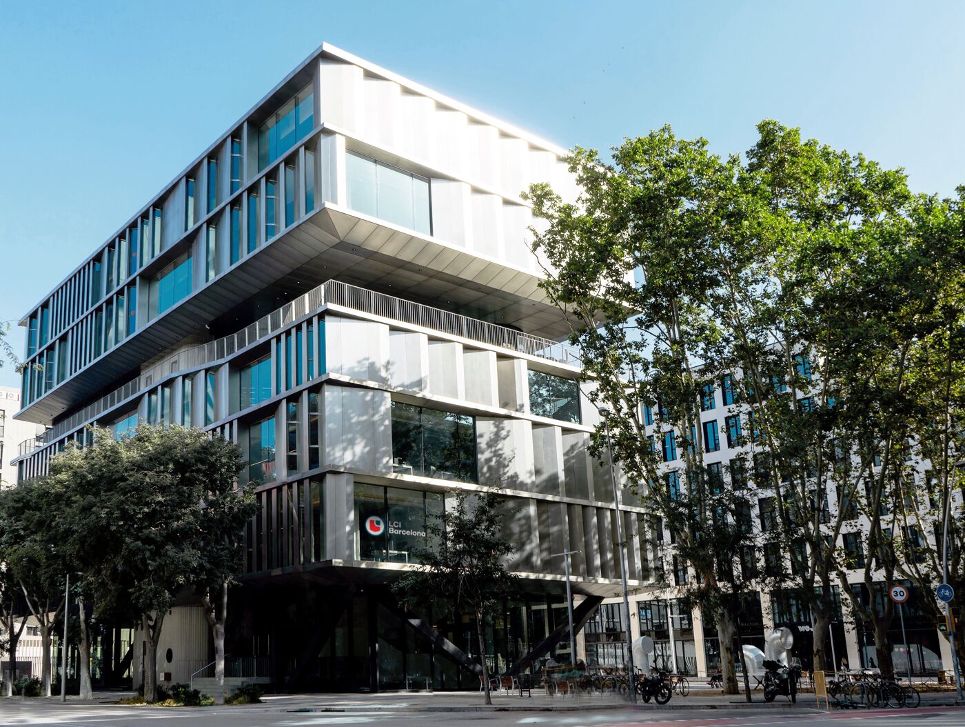 A modern, multi-story building with a unique architectural design stands prominently, partially obscured by lush green trees in front of a slightly less modern, adjacent building.