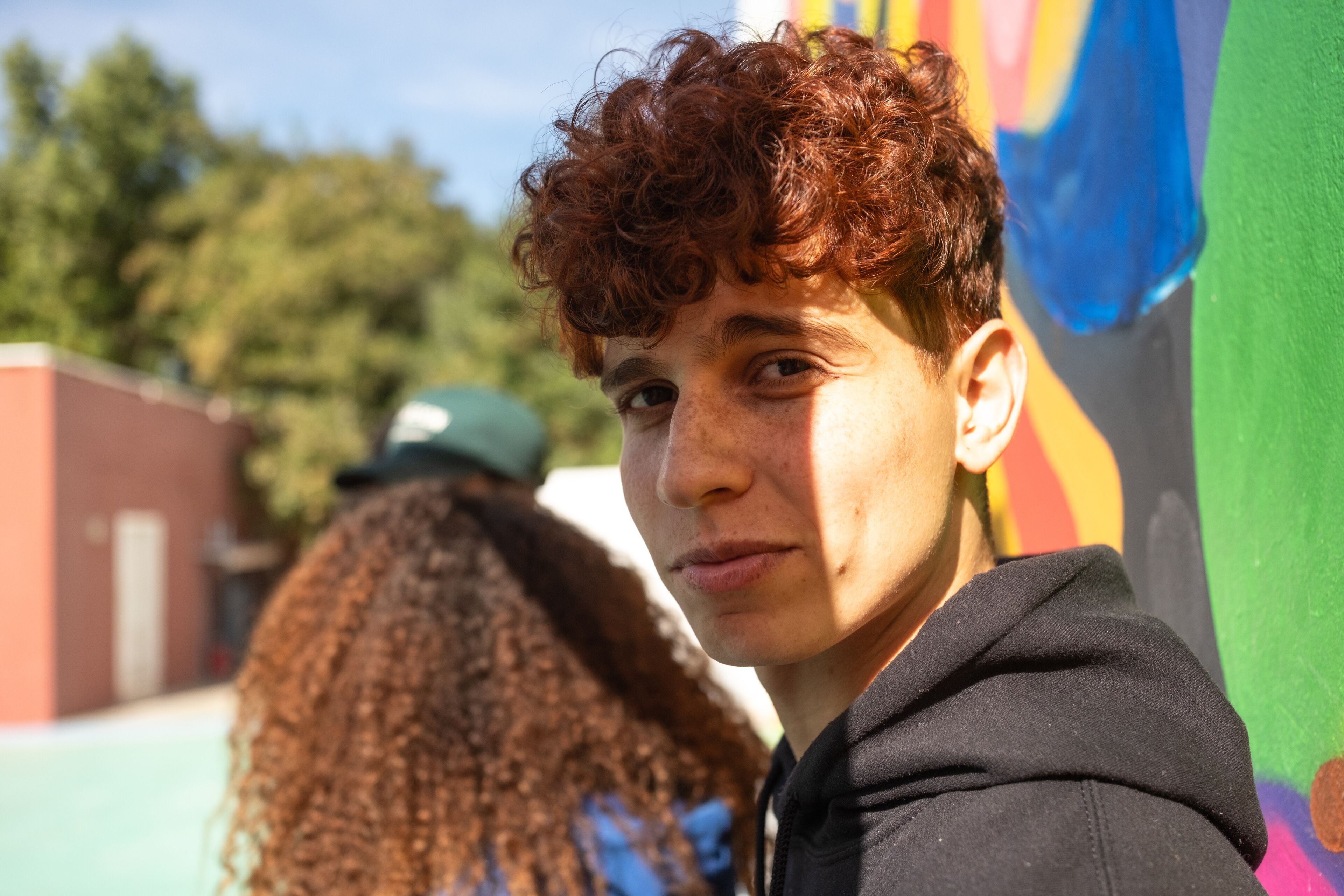 A young individual with curly hair looks contemplative against a vibrant mural backdrop.