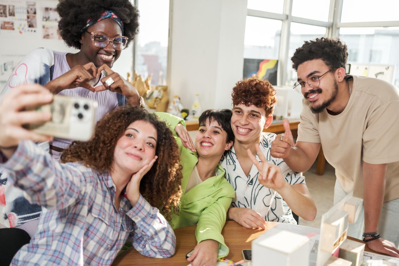 A diverse group of friends poses joyfully for a selfie in a bright and creative indoor space. They express various playful gestures, including heart signs and peace signs, creating a lively and positive atmosphere.