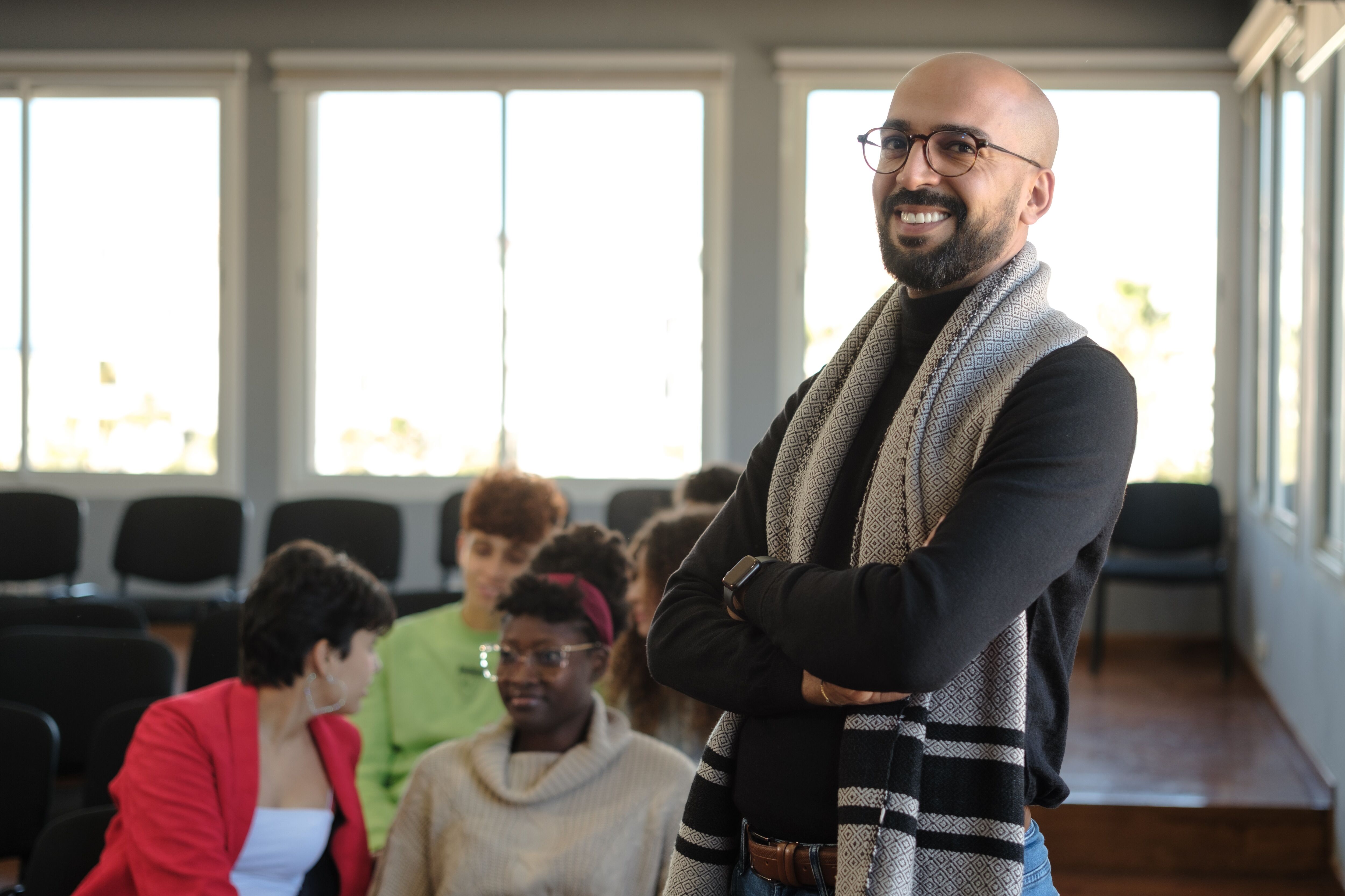 Smiling teacher with crossed arms in a classroom, students in the background.