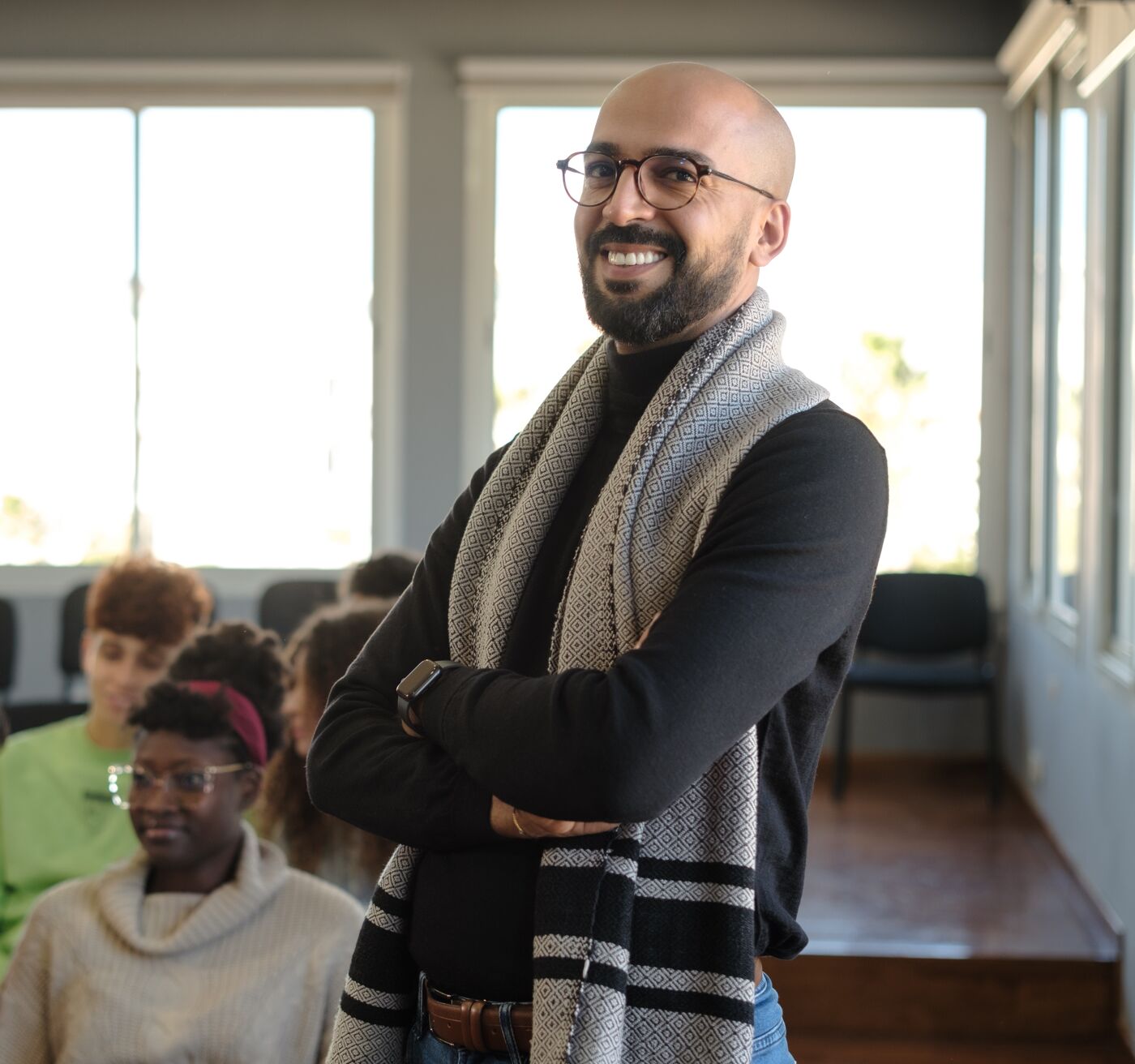 Smiling teacher with crossed arms in a classroom, students in the background.