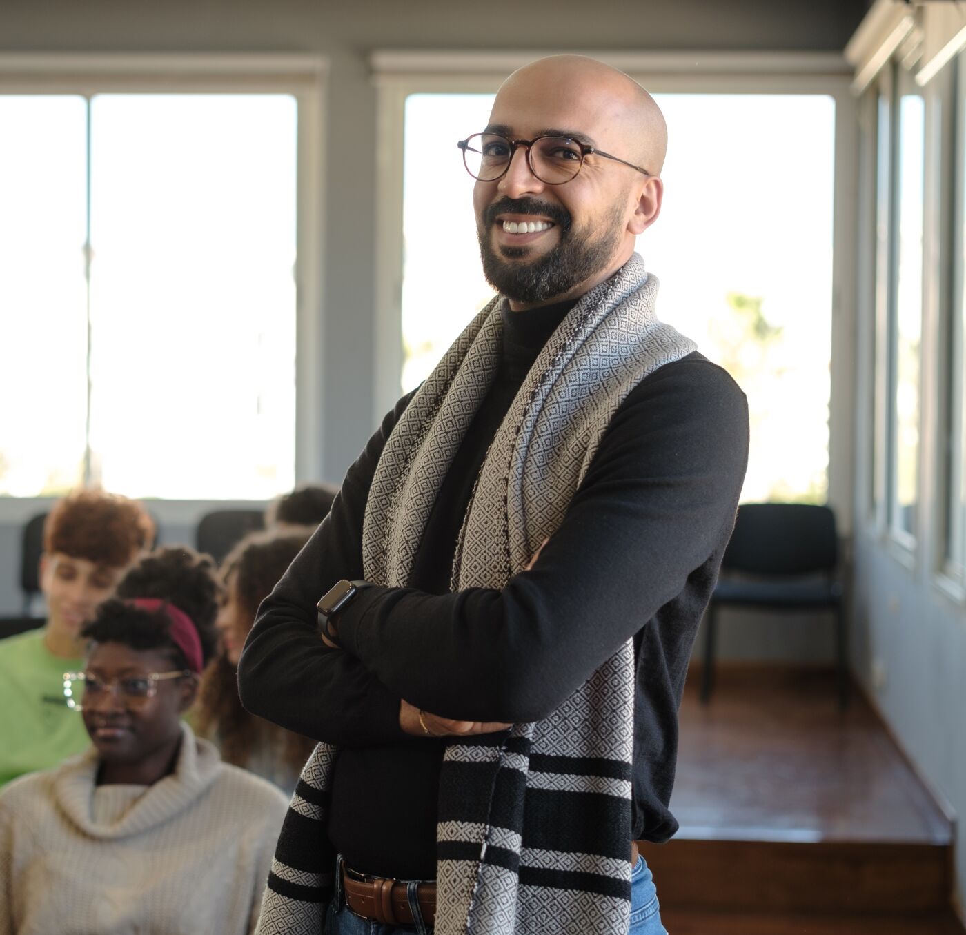 Smiling teacher with crossed arms in a classroom, students in the background.