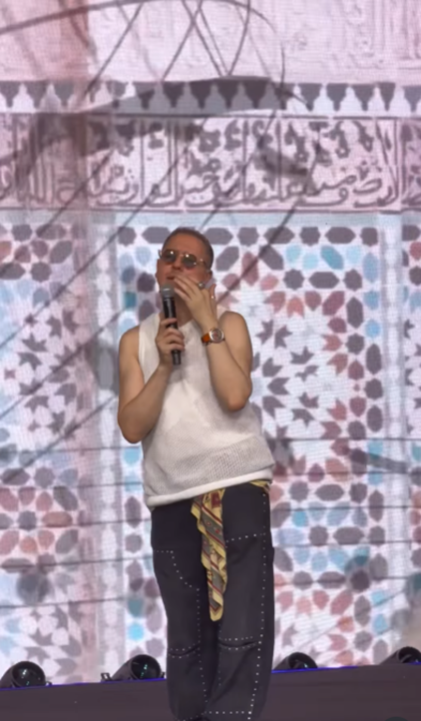 A man with short hair and glasses is holding a microphone and speaking on a stage. He is wearing a white tank top, dark pants, and a patterned scarf around his waist. The background features intricate geometric patterns and Arabic calligraphy.