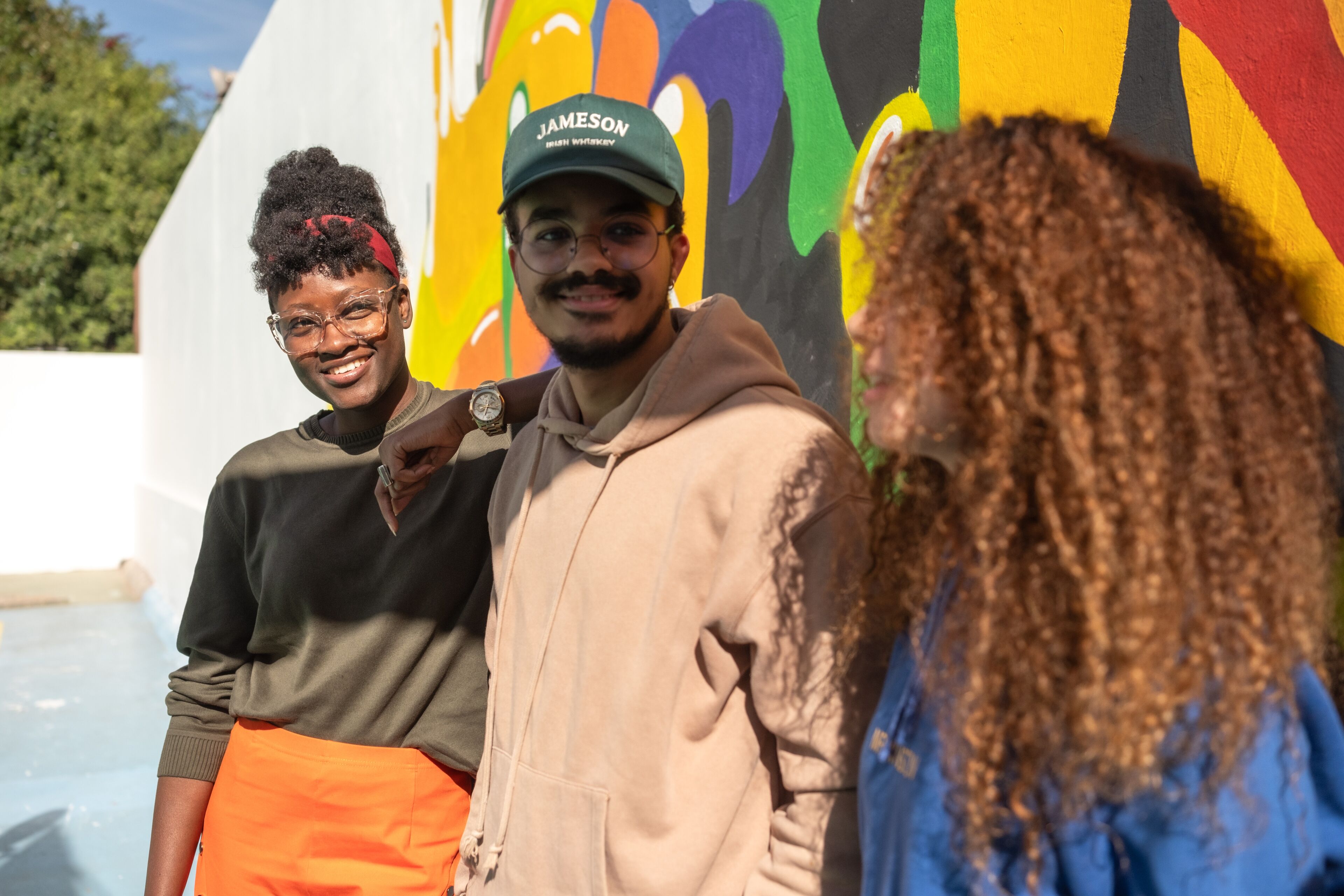 Three friends smile brightly in the sunshine against the backdrop of a colorful mural.