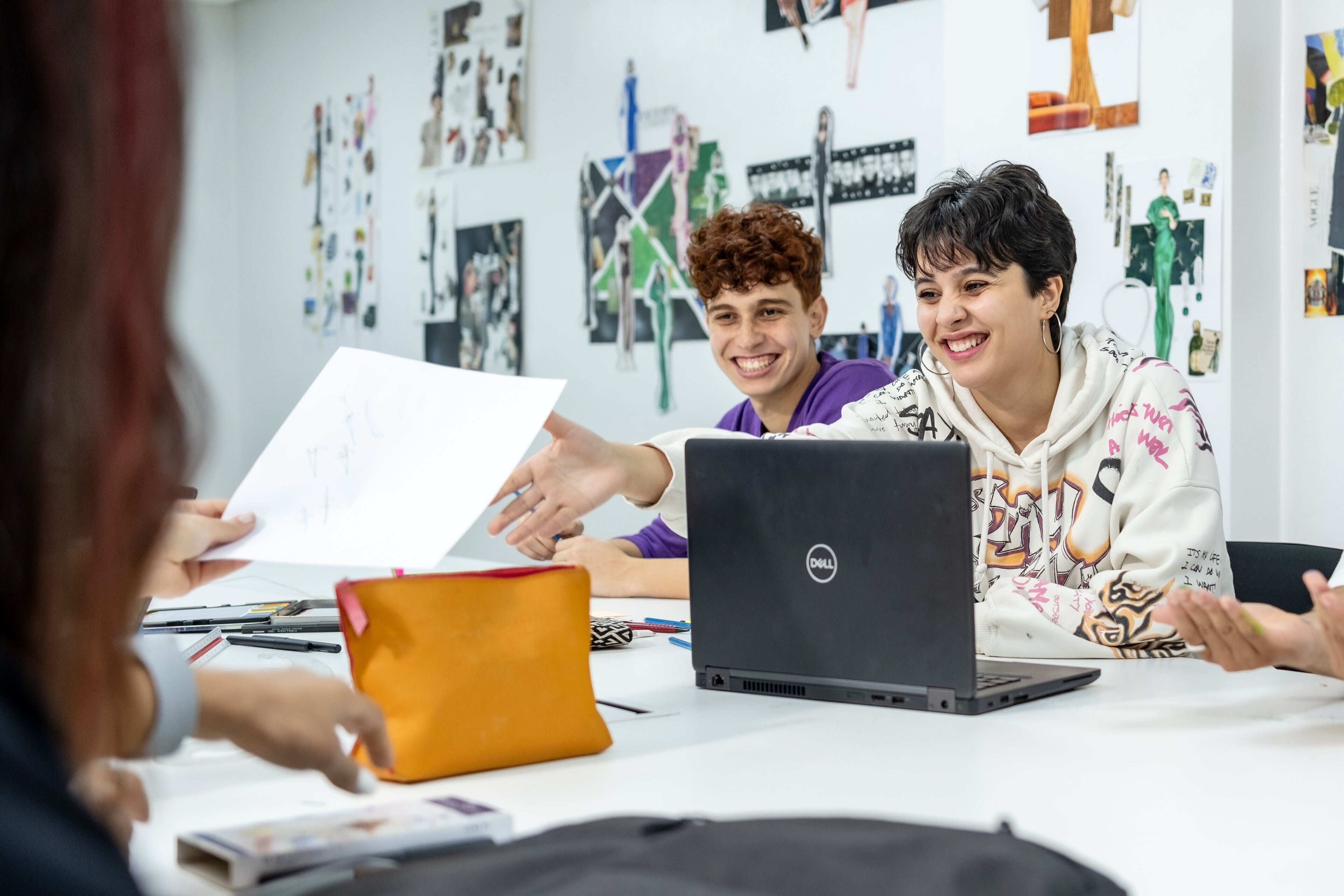 Two young adults engaging with peers in a vibrant creative workshop, sharing sketches while using a laptop.