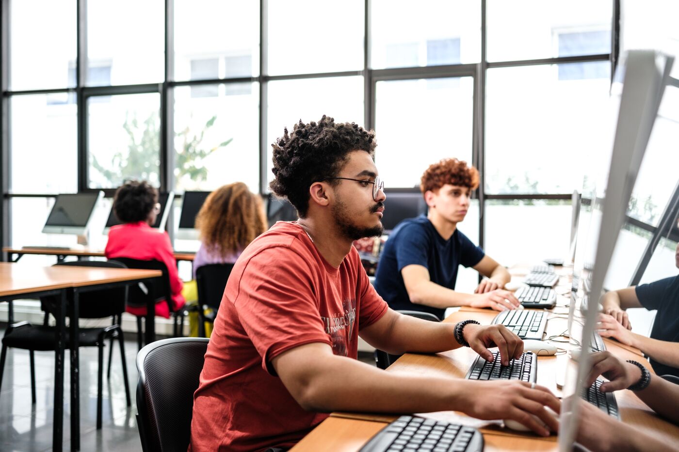 Focused students working on desktop computers in a well-lit computer lab with a view of the outdoors.