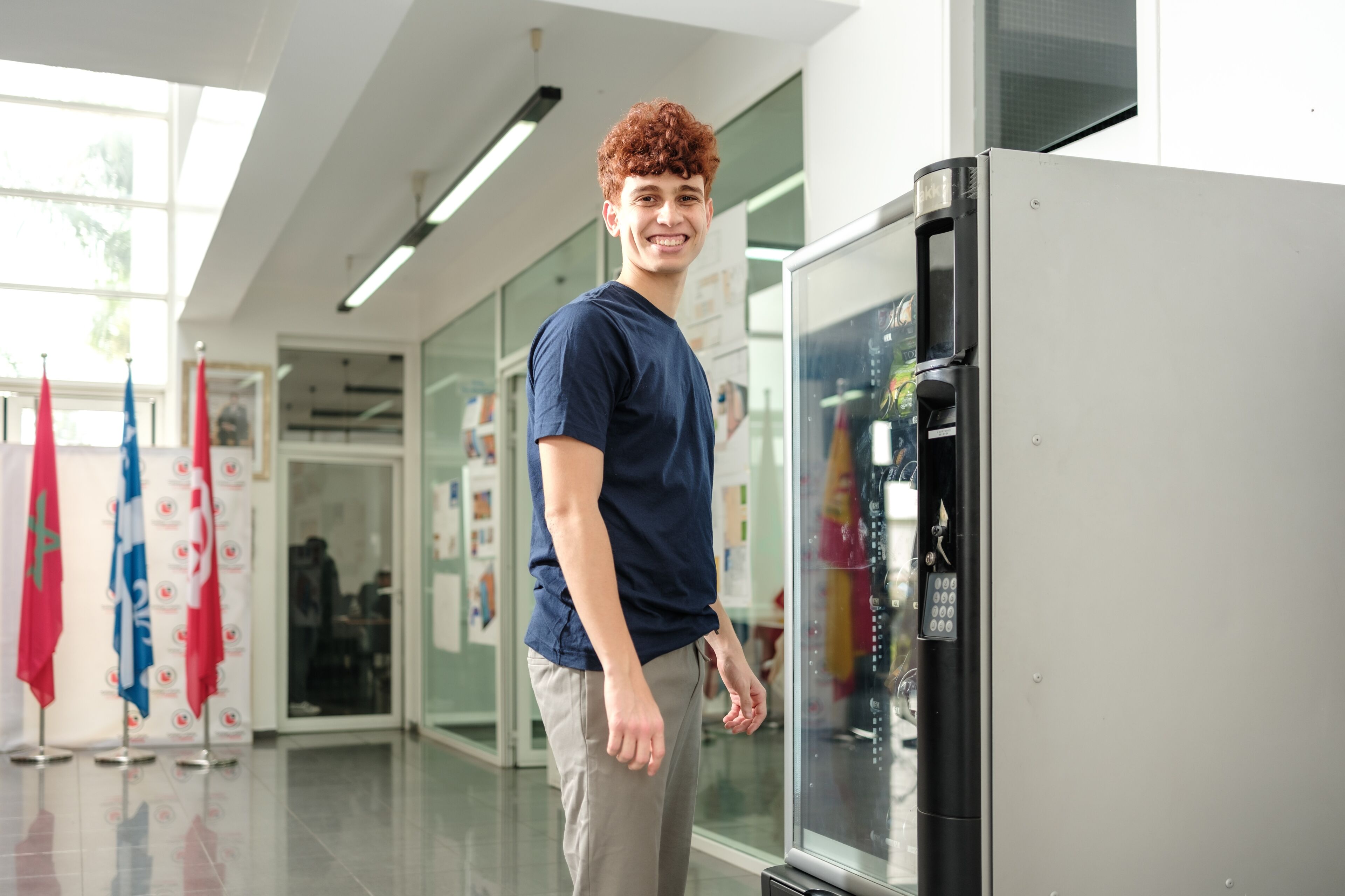 A young man smiles next to a vending machine in a bright corridor of an educational institution.