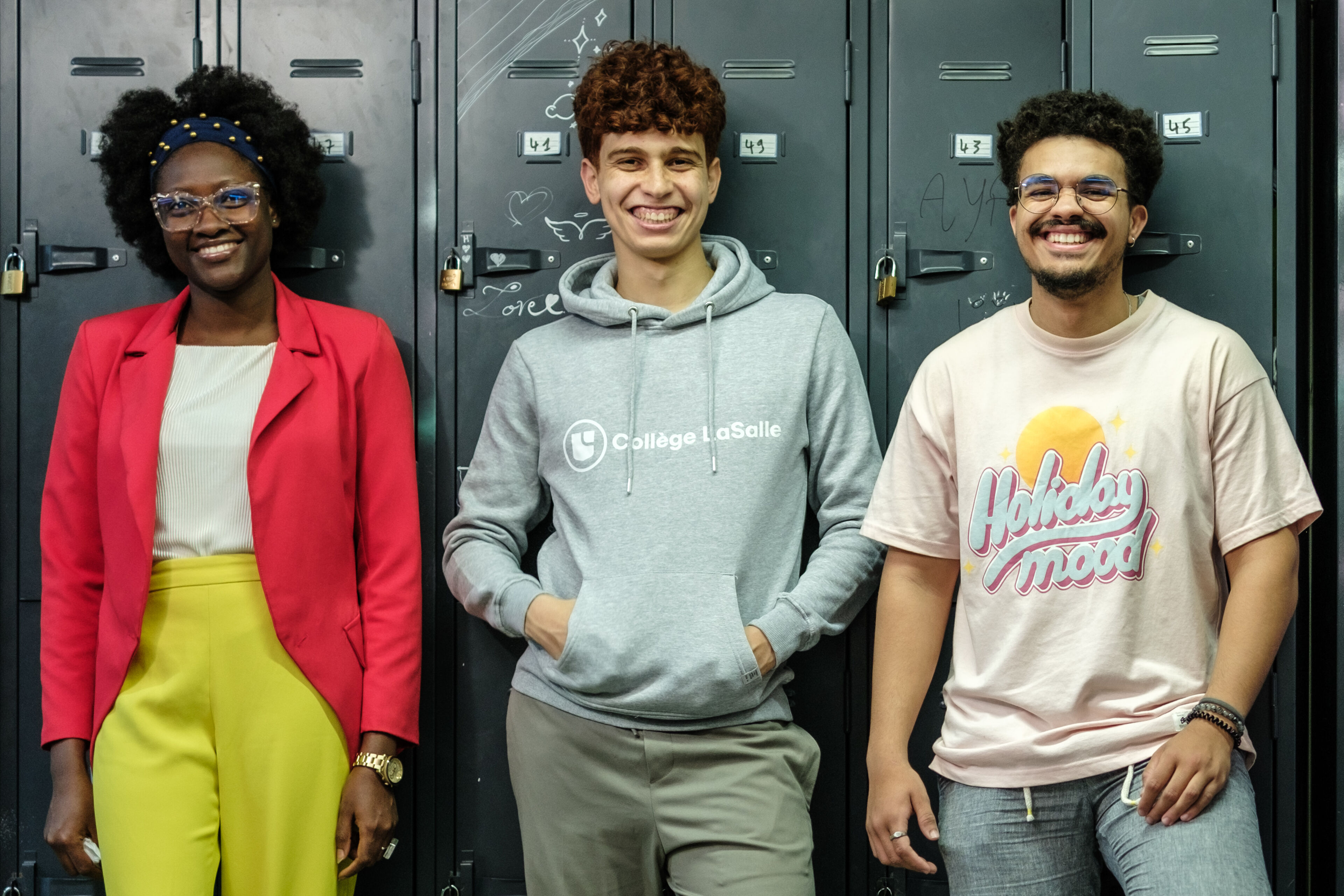 Three students stand smiling in front of their lockers, capturing a moment of campus camaraderie.