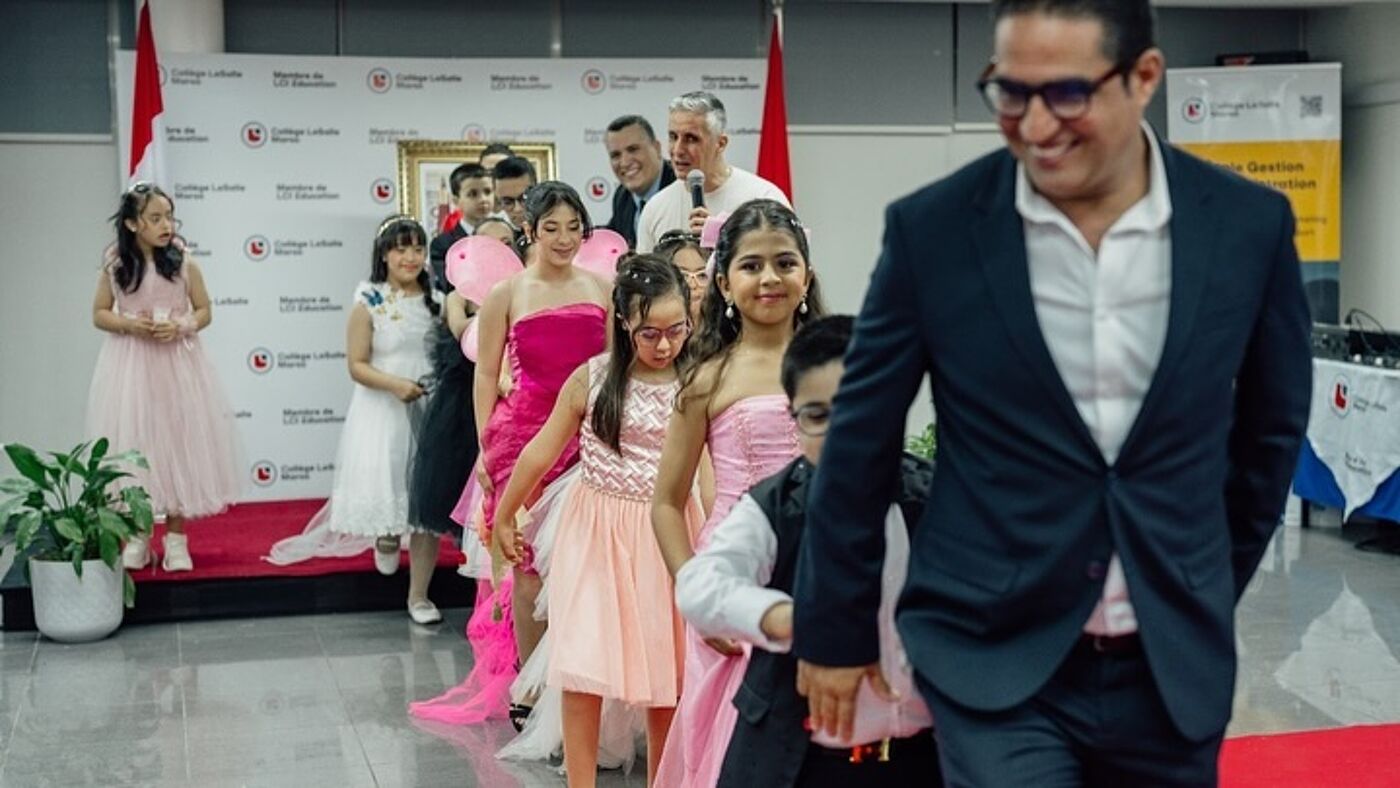 The image shows a group of young girls dressed in pink, walking down a hallway, with a man in a suit and tie following them.