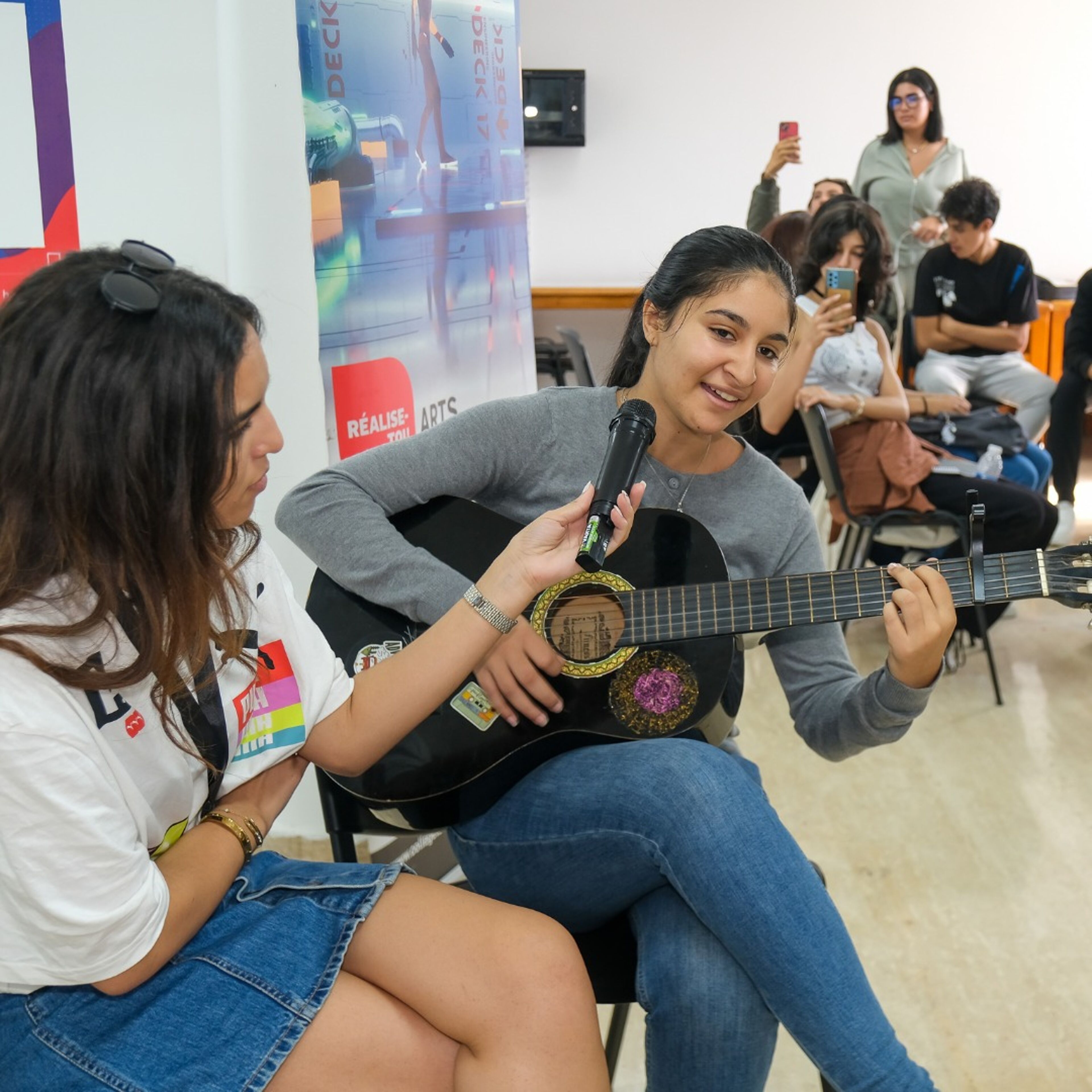  A young woman plays the guitar while smiling and singing, holding a microphone as she performs. Another woman beside her holds the microphone stand, supporting her performance. In the background, a small audience observes, some capturing the moment on their phones.