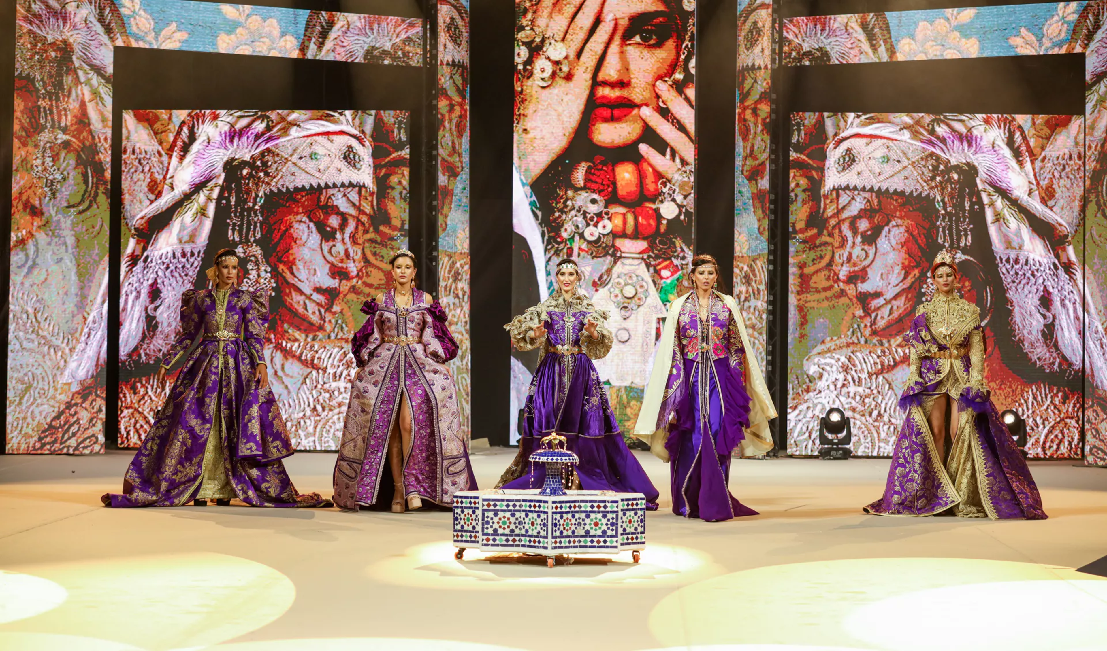 Five models wear luxurious, traditional-inspired purple and gold gowns on a runway, accompanied by intricate jewelry. The backdrop features vibrant cultural artwork, enhancing the rich, ornate theme of the fashion show. A decorative table stands at the center stage.