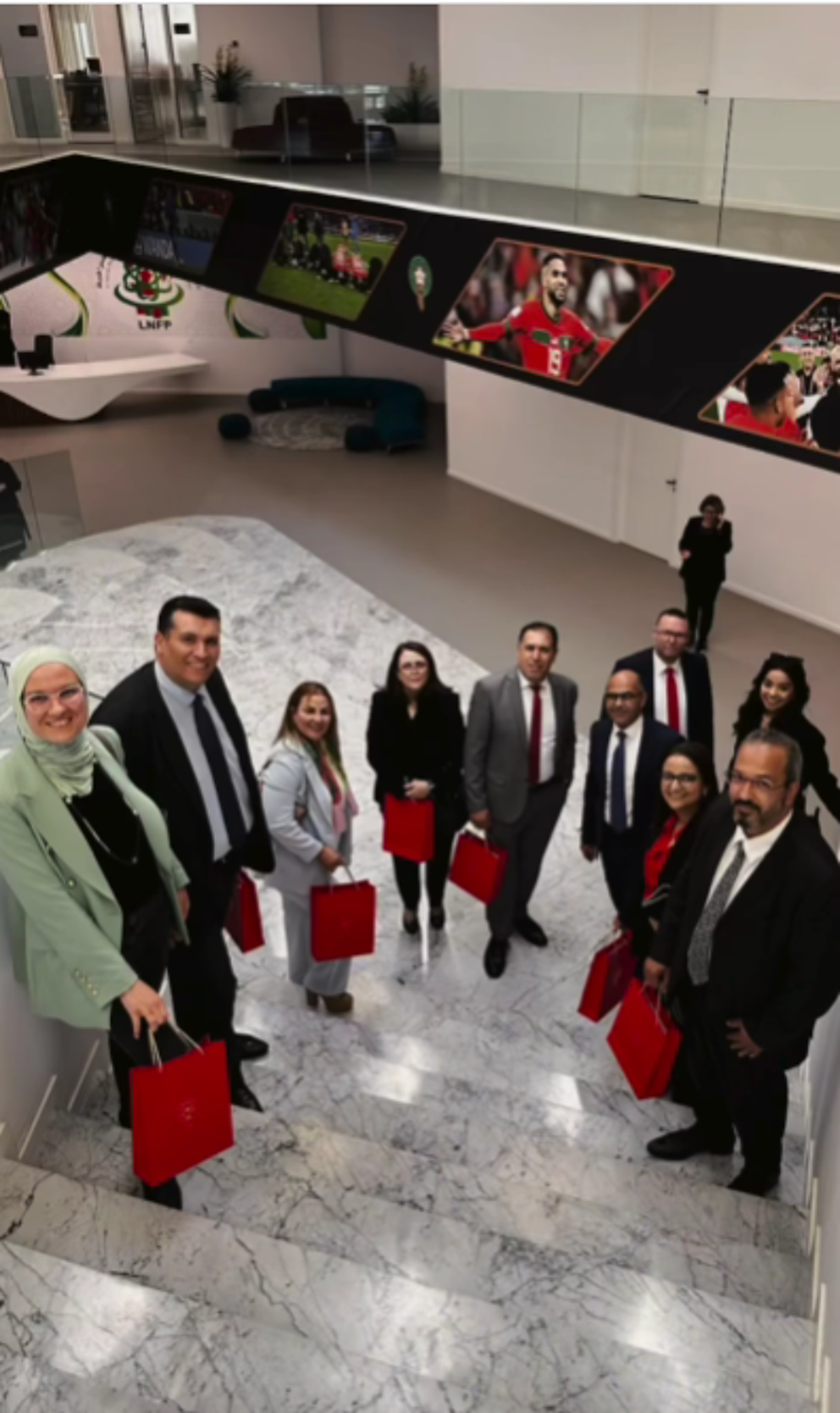 A professional group of men and women stands on a marble staircase inside a modern building, holding red gift bags. Sports-themed decorations, including framed images of athletes, adorn the interior walls, creating a dynamic and vibrant atmosphere.