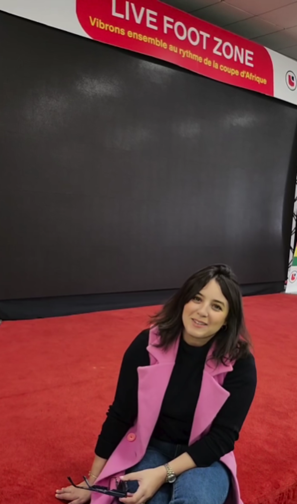 A woman sits casually on a red carpet in front of a large black screen at the "Live Foot Zone," with the banner reading, "Vibrons ensemble au rythme de la coupe d'Afrique" (Let's vibrate to the rhythm of the African Cup). The space is designed for a vibrant football event atmosphere.