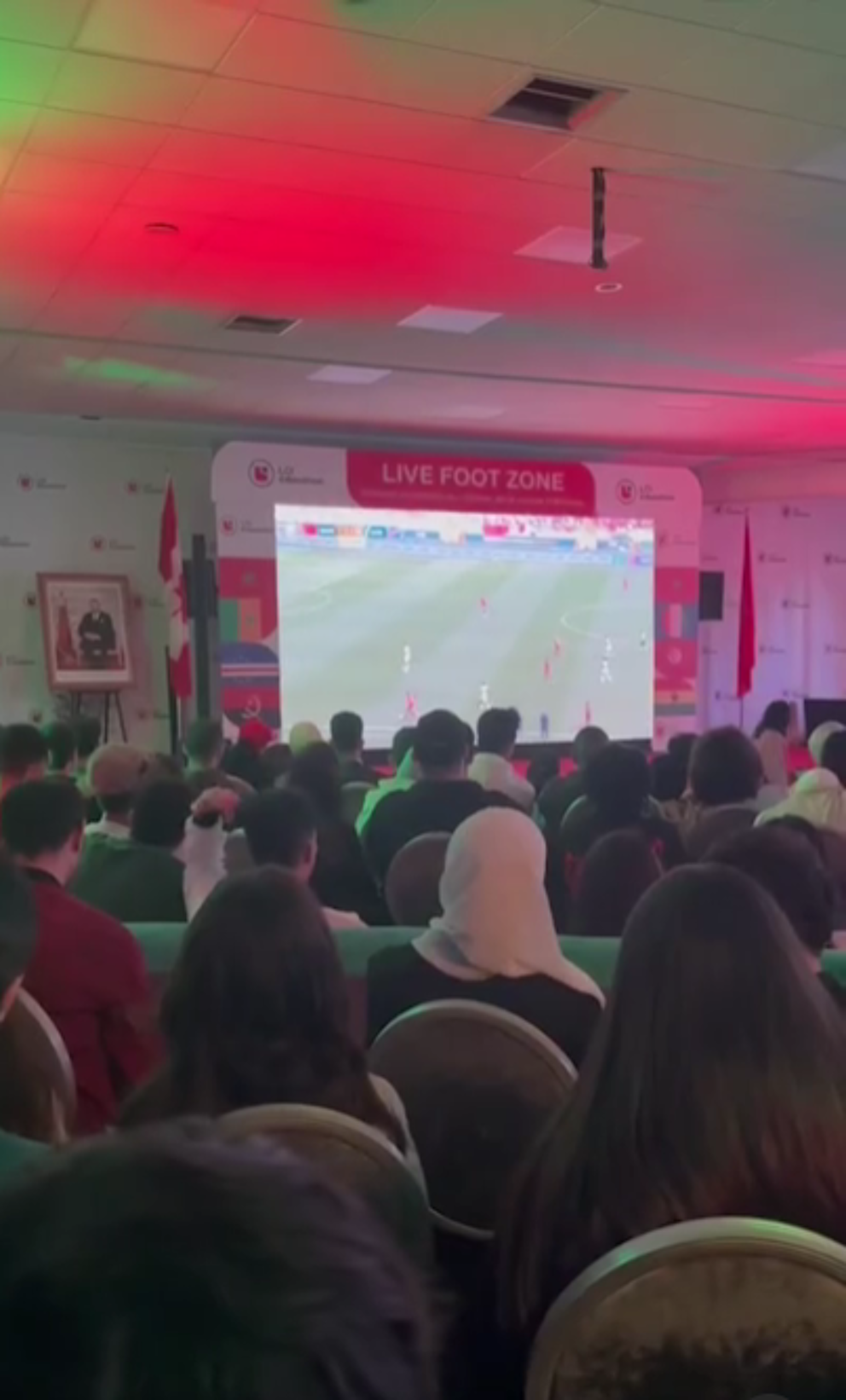 The image shows a large crowd of people sitting in chairs, watching a soccer game on a big screen TV.