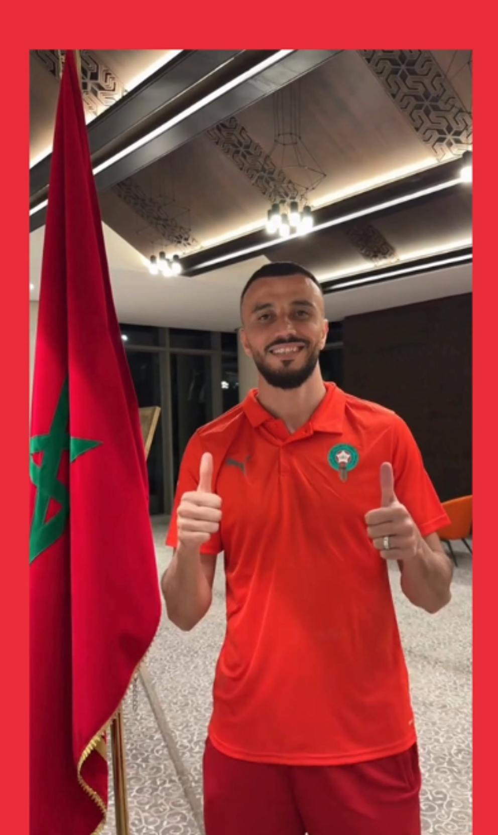 The picture features a man wearing a red and orange soccer jersey, giving a thumbs-up sign in front of a flag.