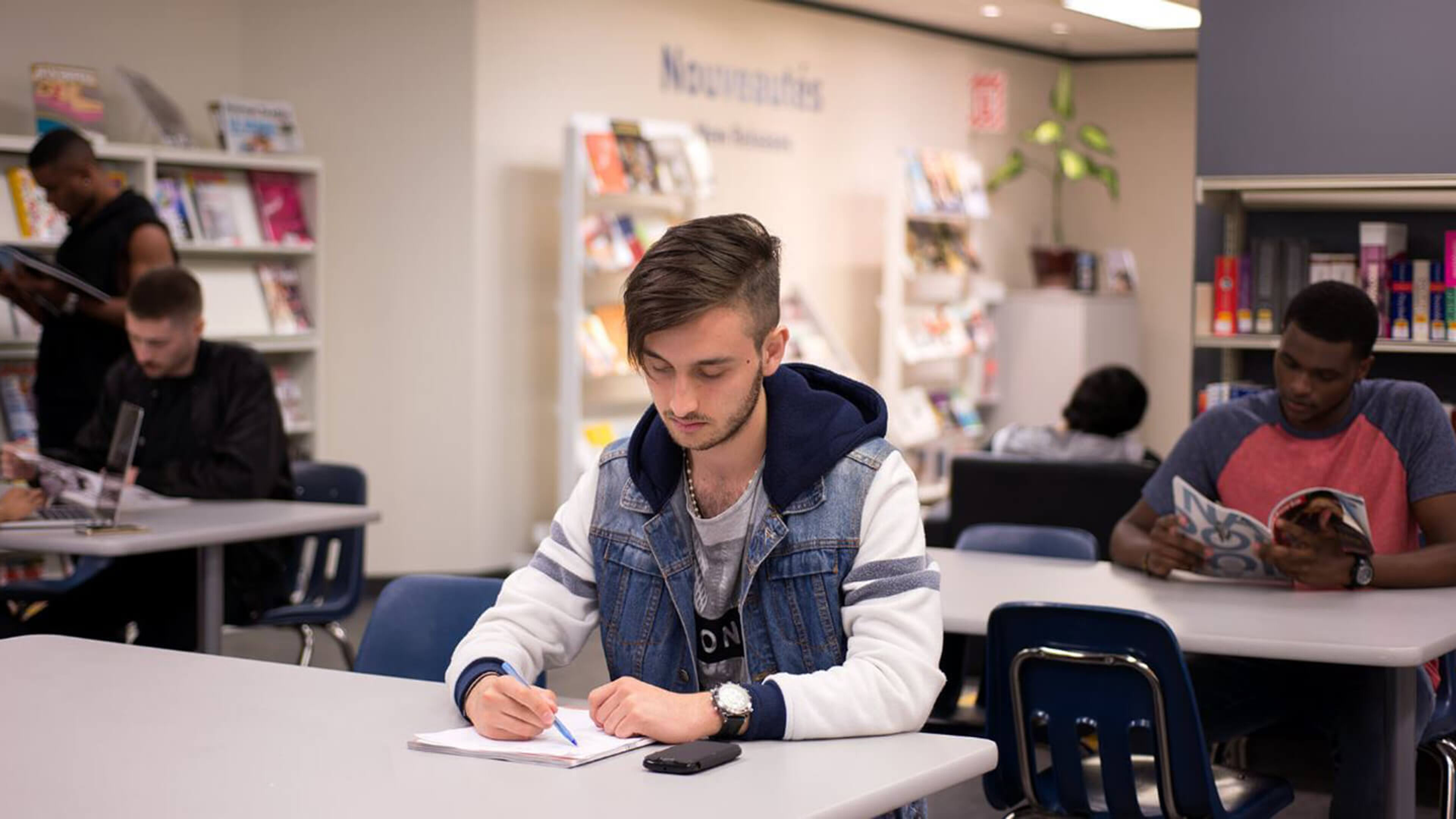 A young man in a denim jacket intently writes notes at a library table, while others read around him.