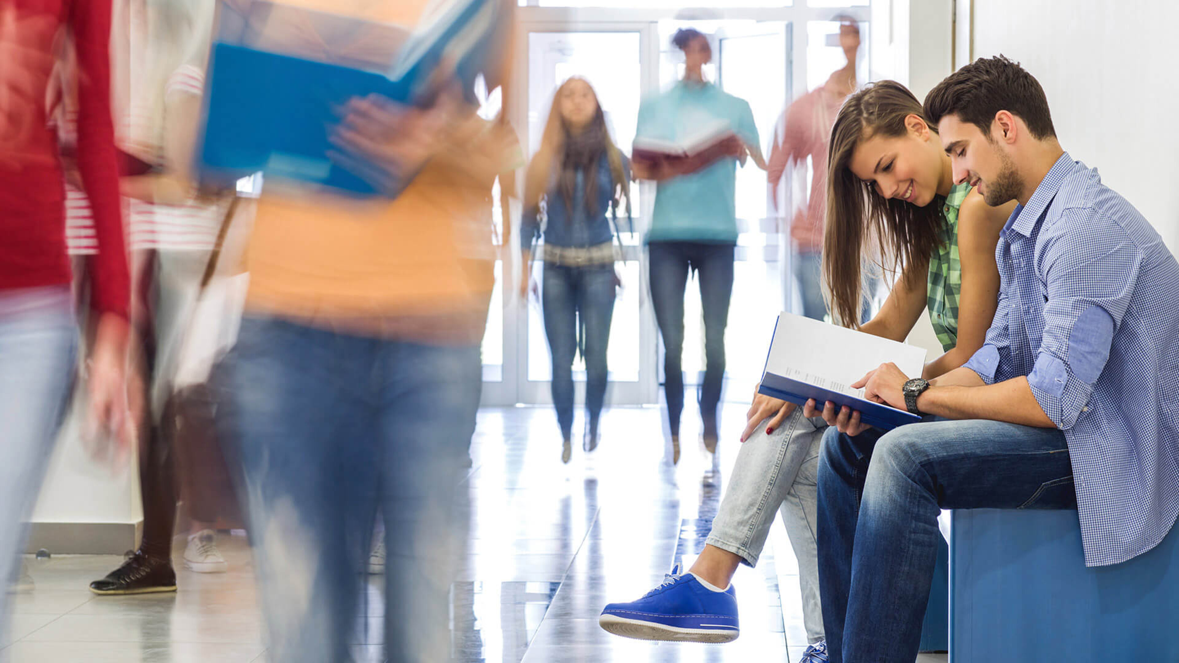 A focused young man and woman review a textbook together on a bench in a bustling school corridor, surrounded by motion-blurred figures of passing students.