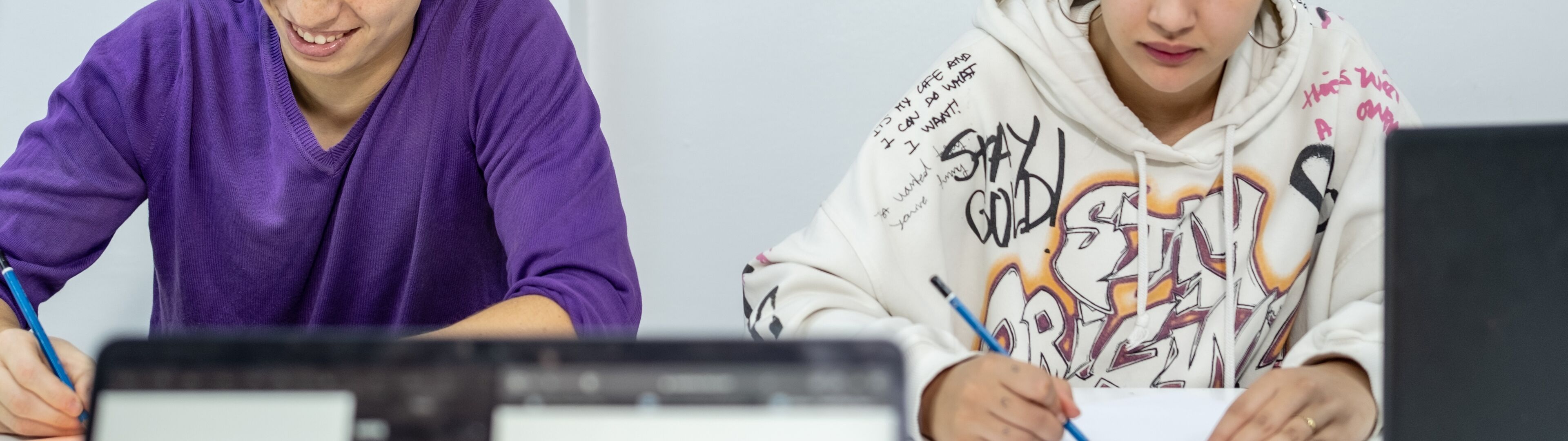 Un garçon et une fille, probablement adolescents, étudient à un bureau encombré de papiers et d'ordinateurs portables, concentrés sur leur travail dans une pièce décorée de posters.

