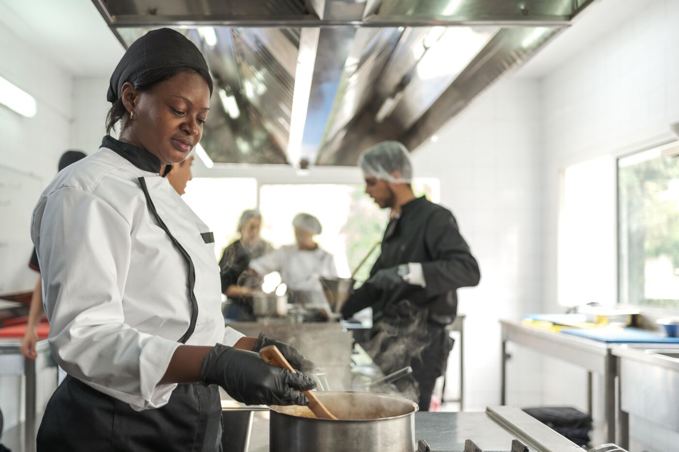  A chef wearing a white uniform and black gloves stirs a pot on the stove in a bustling kitchen. Other chefs, dressed in black and white uniforms, work in the background, showcasing teamwork and culinary expertise in a clean, well-equipped space.