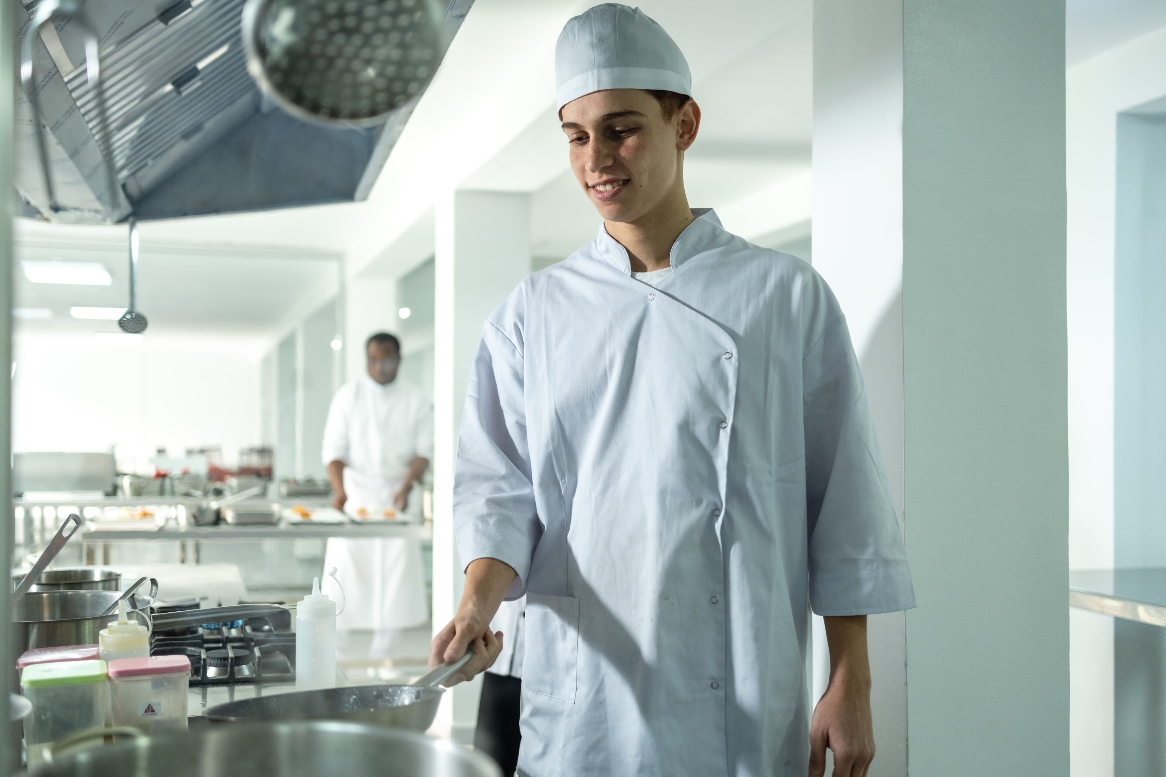 Un jeune chef en uniforme blanc sourit en cuisinant dans une cuisine professionnelle lumineuse, avec un collègue travaillant en arrière-plan.