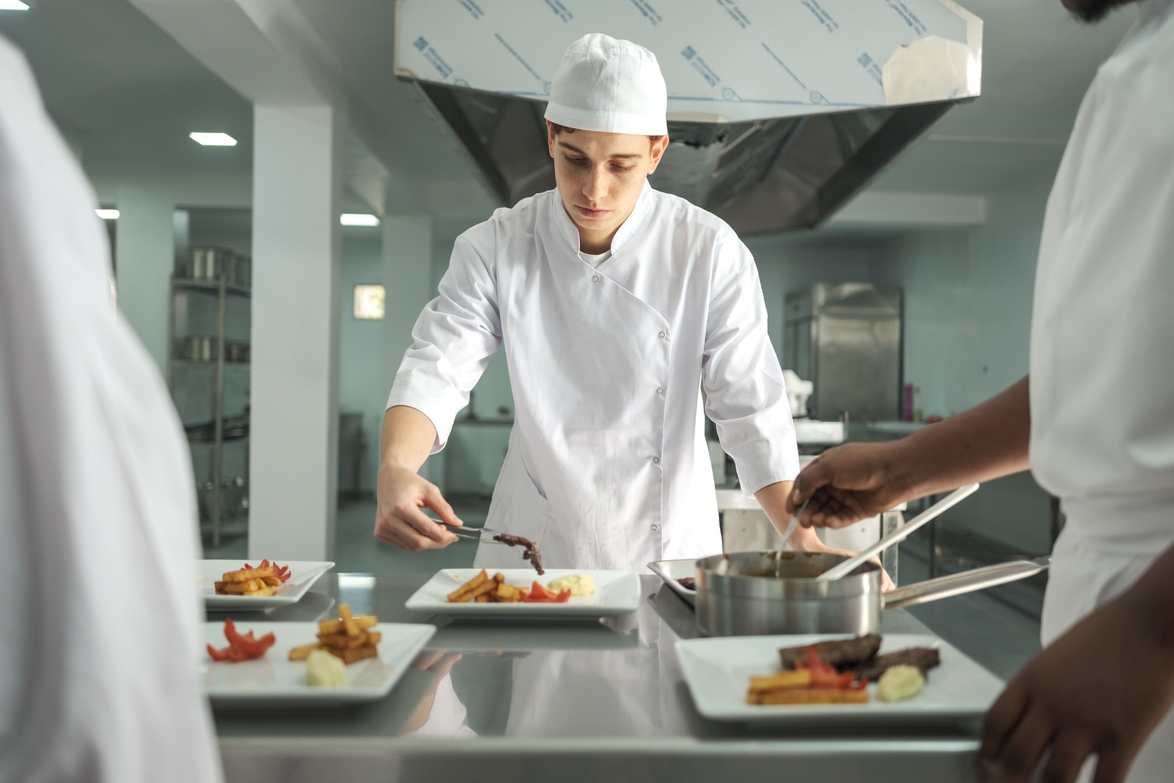Jeune chef concentré en uniforme blanc arrangeant méticuleusement les plats dans une cuisine professionnelle, collègues à ses côtés.