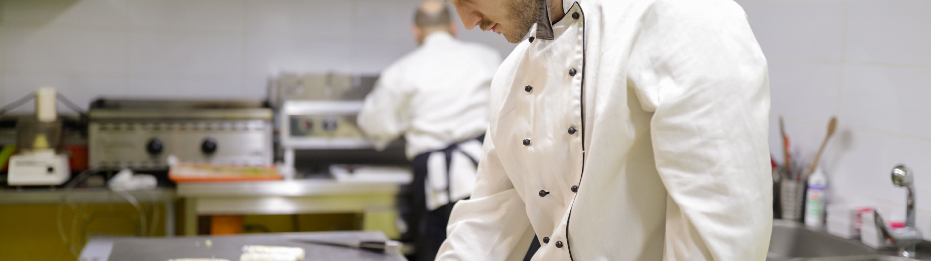 Un chef concentré en tenue traditionnelle blanche pétrit de la pâte sur un comptoir en acier inoxydable, avec un collègue en arrière-plan.