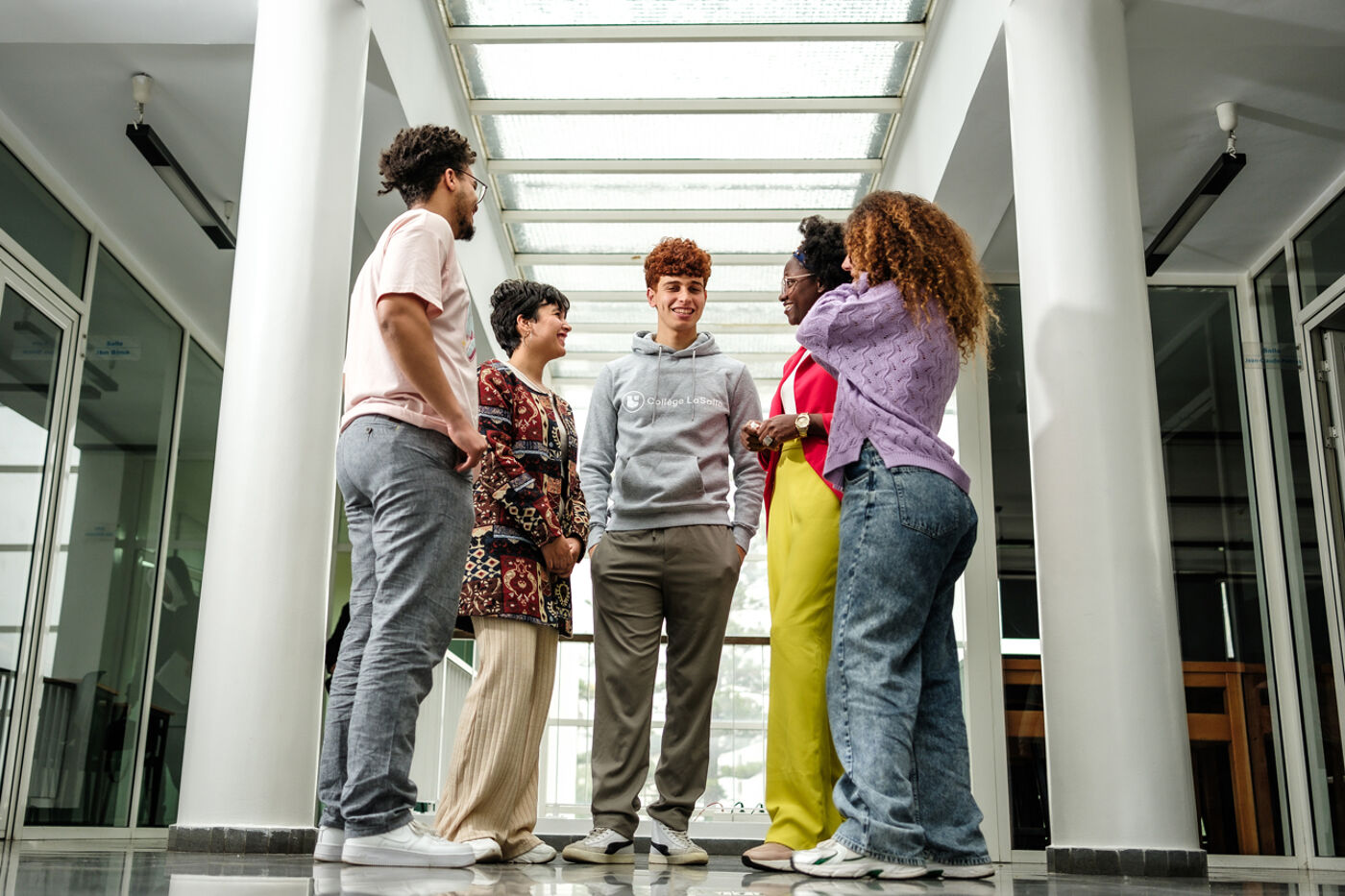 Five young adults are engaged in a friendly chat, standing in a semi-circle inside a bright corridor with a translucent ceiling.