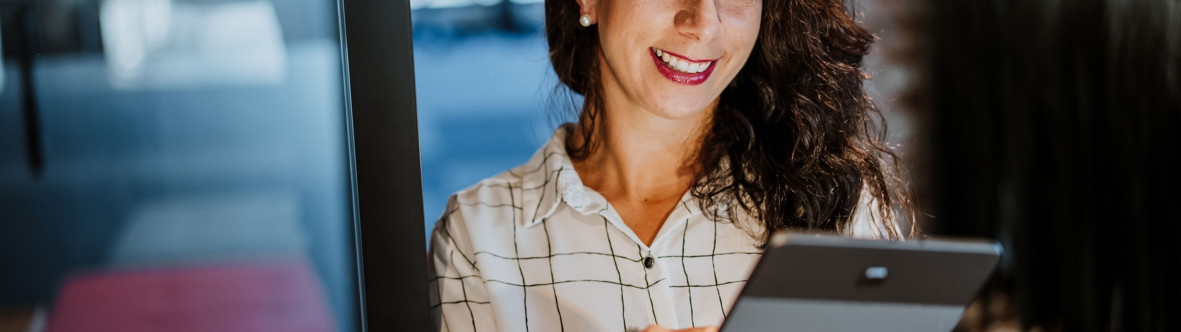 Une femme d'affaires confiante en chemise à carreaux blanche sourit en tenant une tablette numérique dans un environnement de bureau.