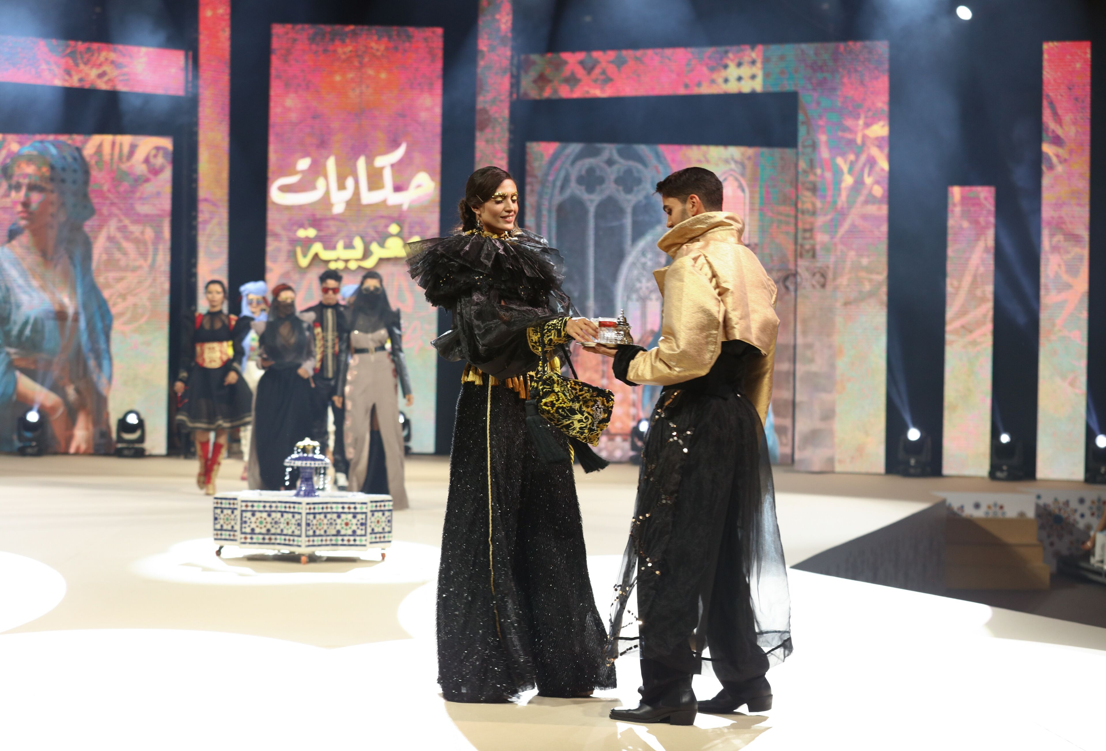 Deux mannequins échangent des boîtes ornées sur le podium, sur fond de défilé de mode animé.