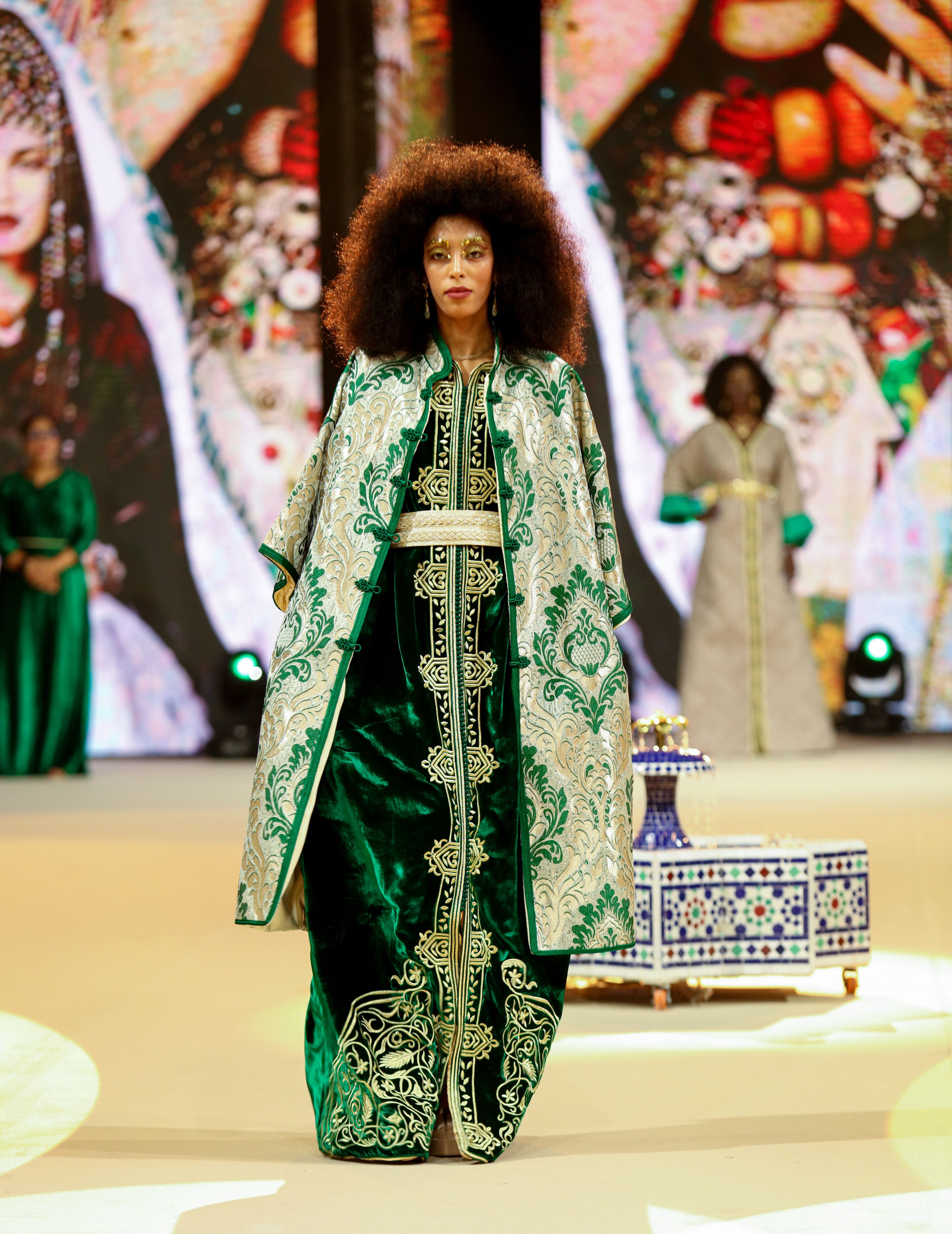 Un mannequin à la coiffure afro présente une élégante robe traditionnelle verte et dorée lors d'un défilé de mode.
