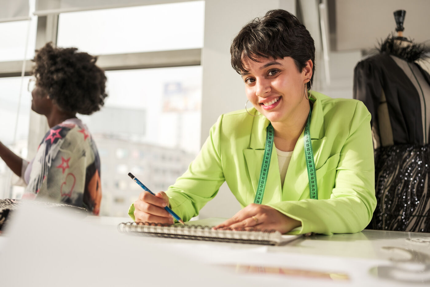A smiling woman with short hair, adorned with hoop earrings, wearing a lime green blazer and a measuring tape around her neck, is typing on a keyboard.

