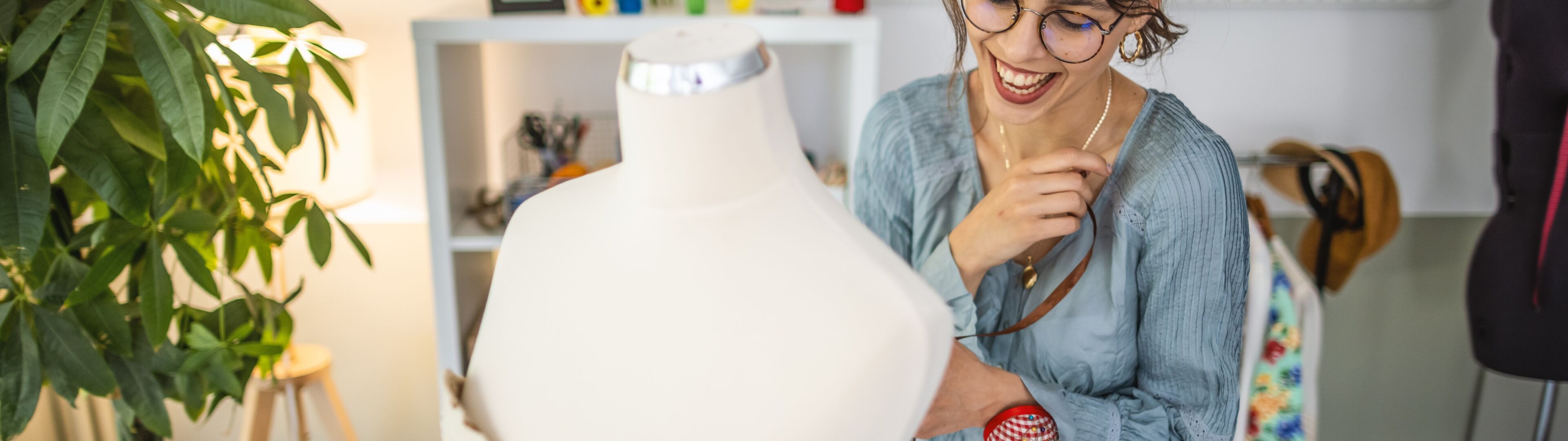Une créatrice de mode souriante ajuste un mannequin dans son atelier lumineux et plein de plantes, entourée d'esquisses de mode.