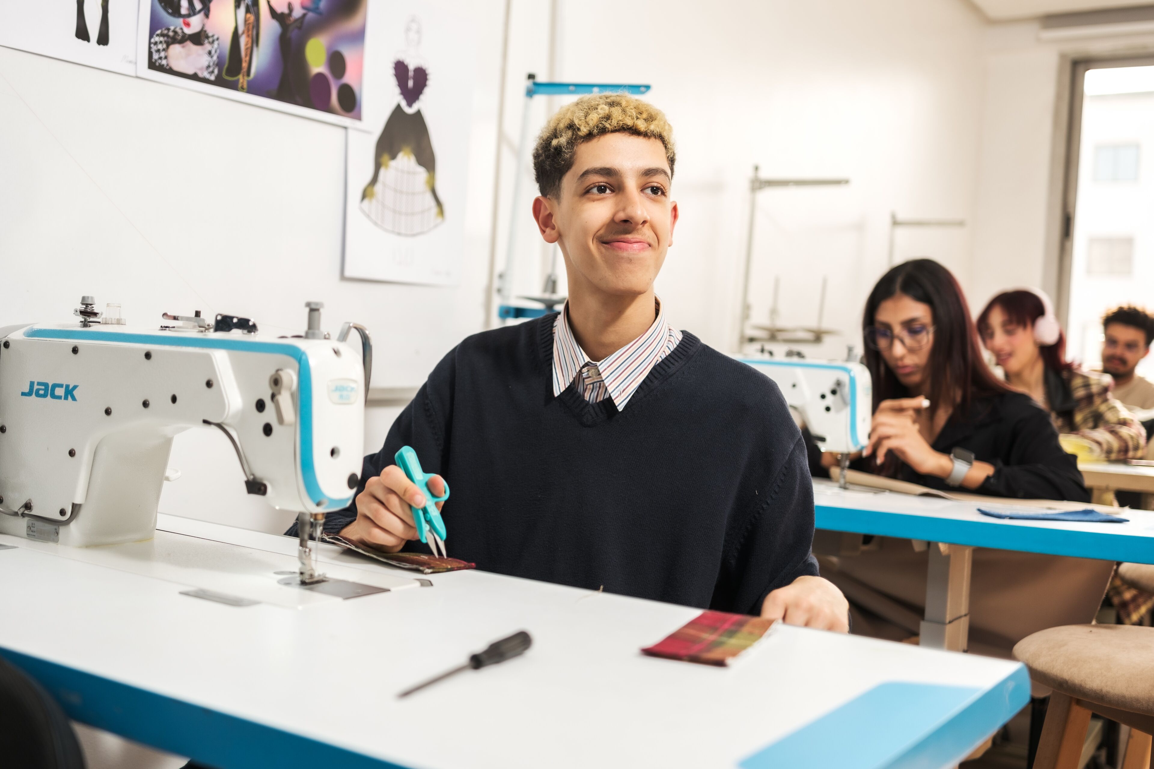 Un jeune étudiant aux cheveux bouclés sourit, tenant des ciseaux dans une classe de design de mode, avec des machines à coudre et des camarades concentrés en arrière-plan.