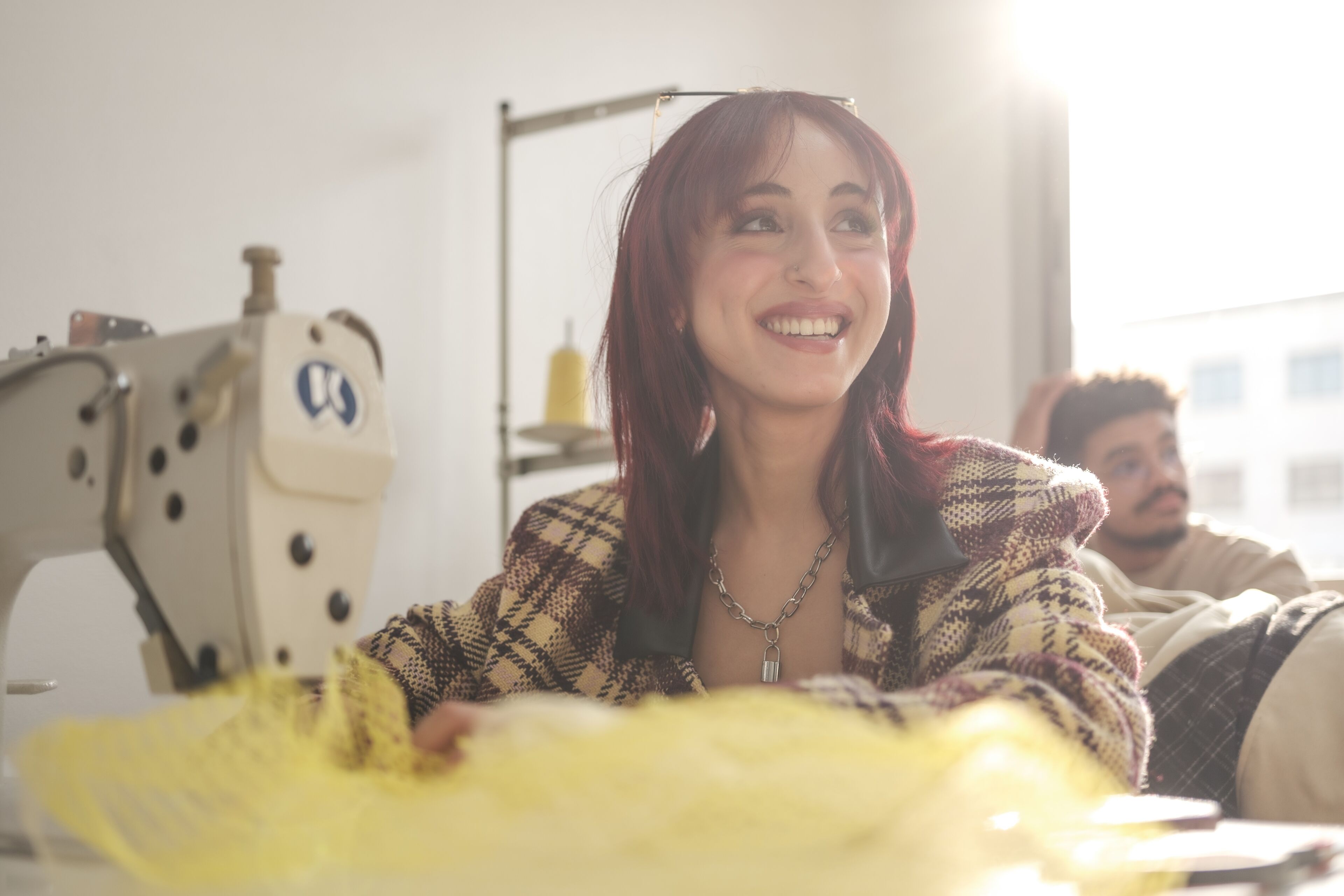 Une femme souriante aux cheveux rouges travaillant sur un tissu jaune avec une machine à coudre, dans un atelier bien éclairé, avec un collègue masculin en arrière-plan.