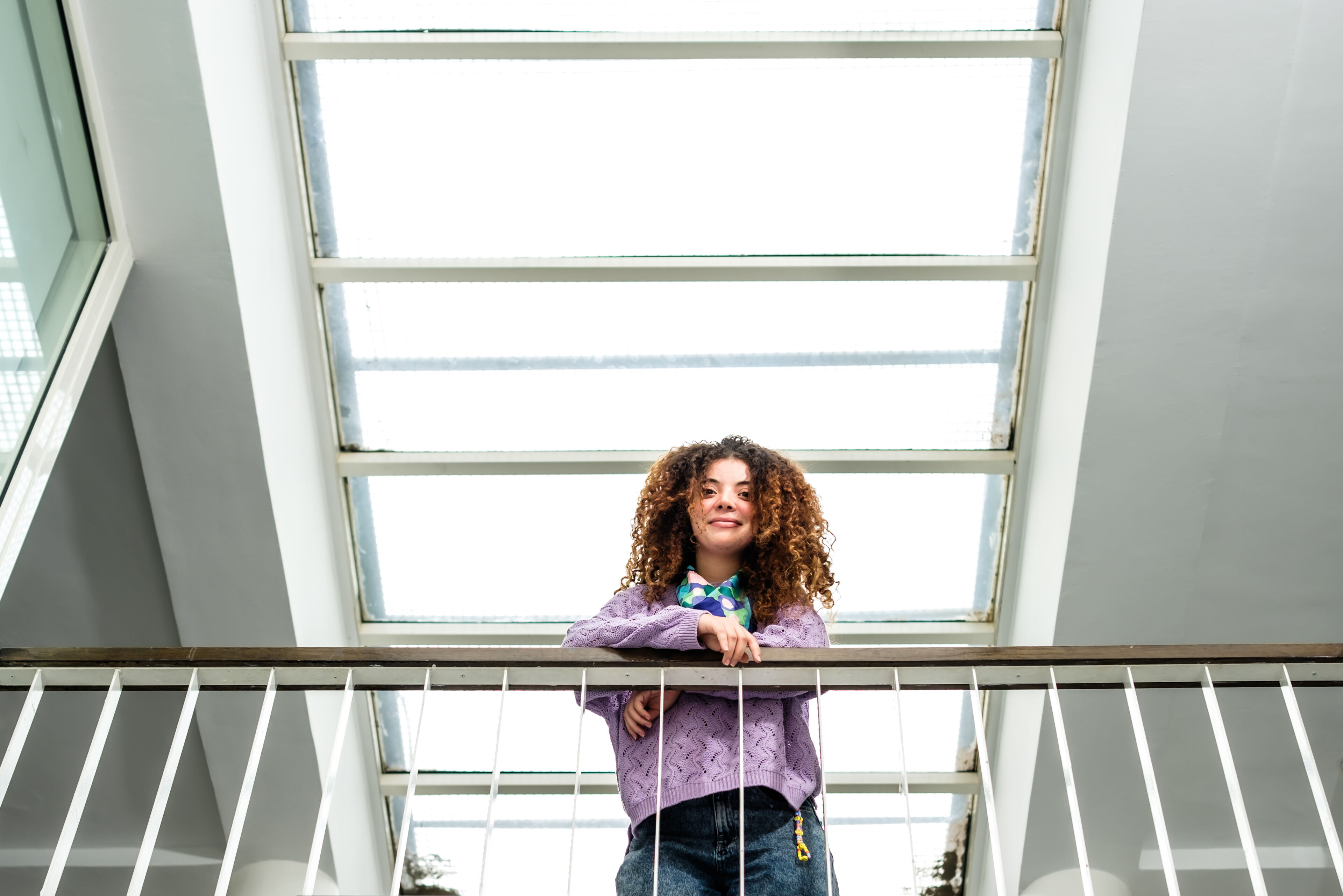 A cheerful woman with curly hair leans on a metal railing, basking in the natural light coming through a large skylight above.