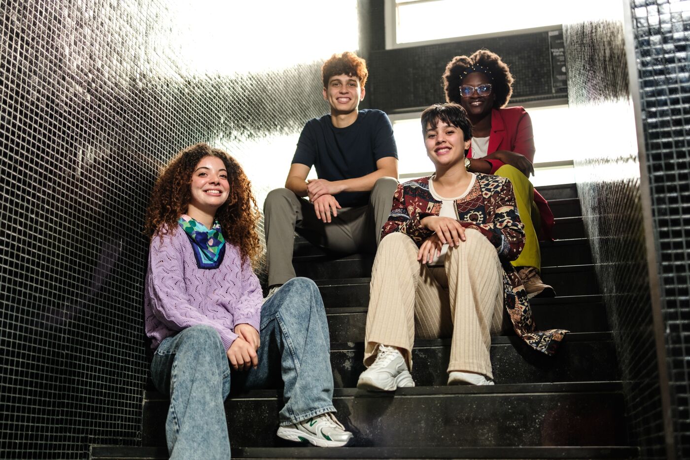 Four young adults, two women in the front and a man and a woman behind, are sitting on dark stairs, smiling and posing for a photo. They are casually dressed, showcasing individual styles.