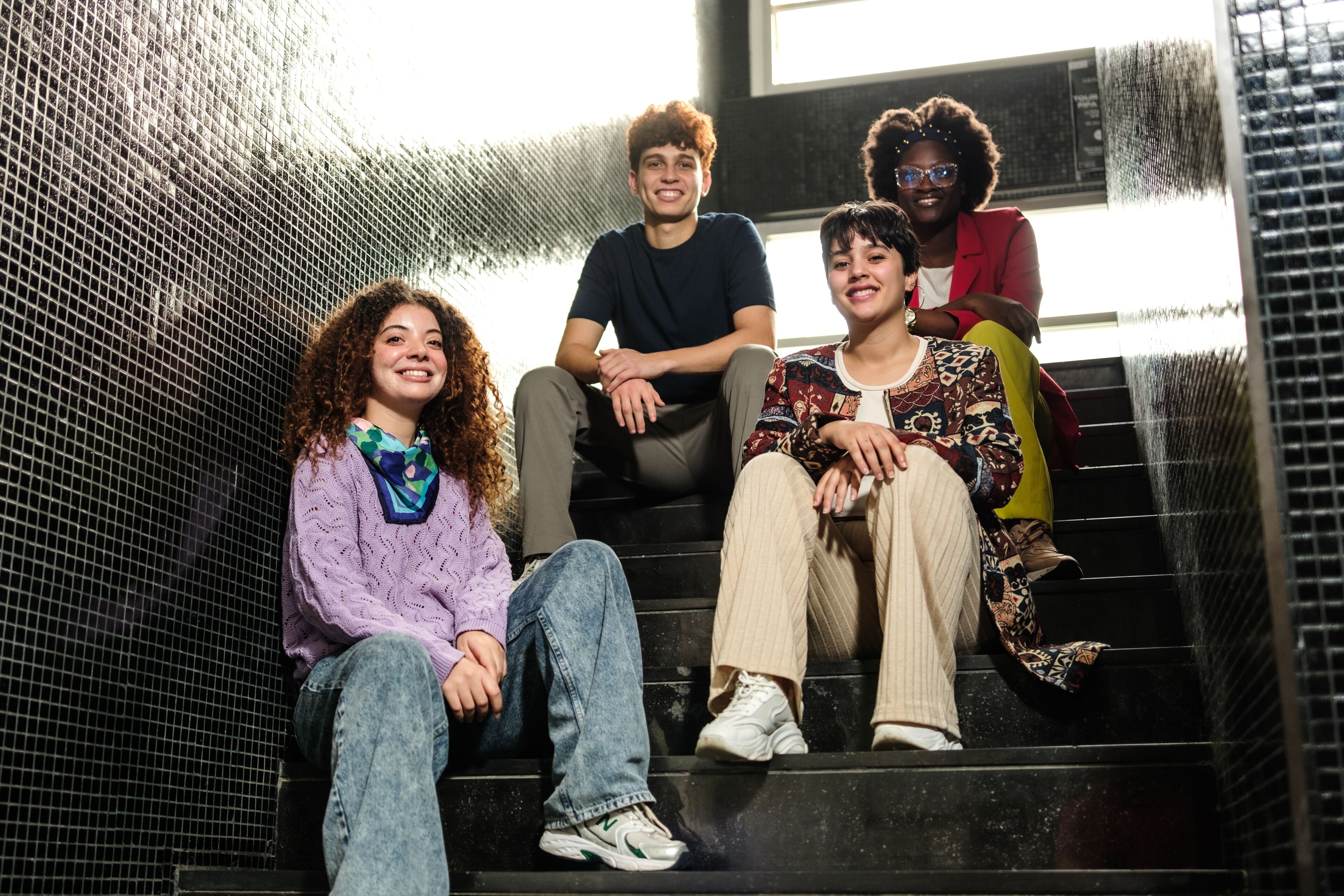 Four young adults, two women in the front and a man and a woman behind, are sitting on dark stairs, smiling and posing for a photo. They are casually dressed, showcasing individual styles.