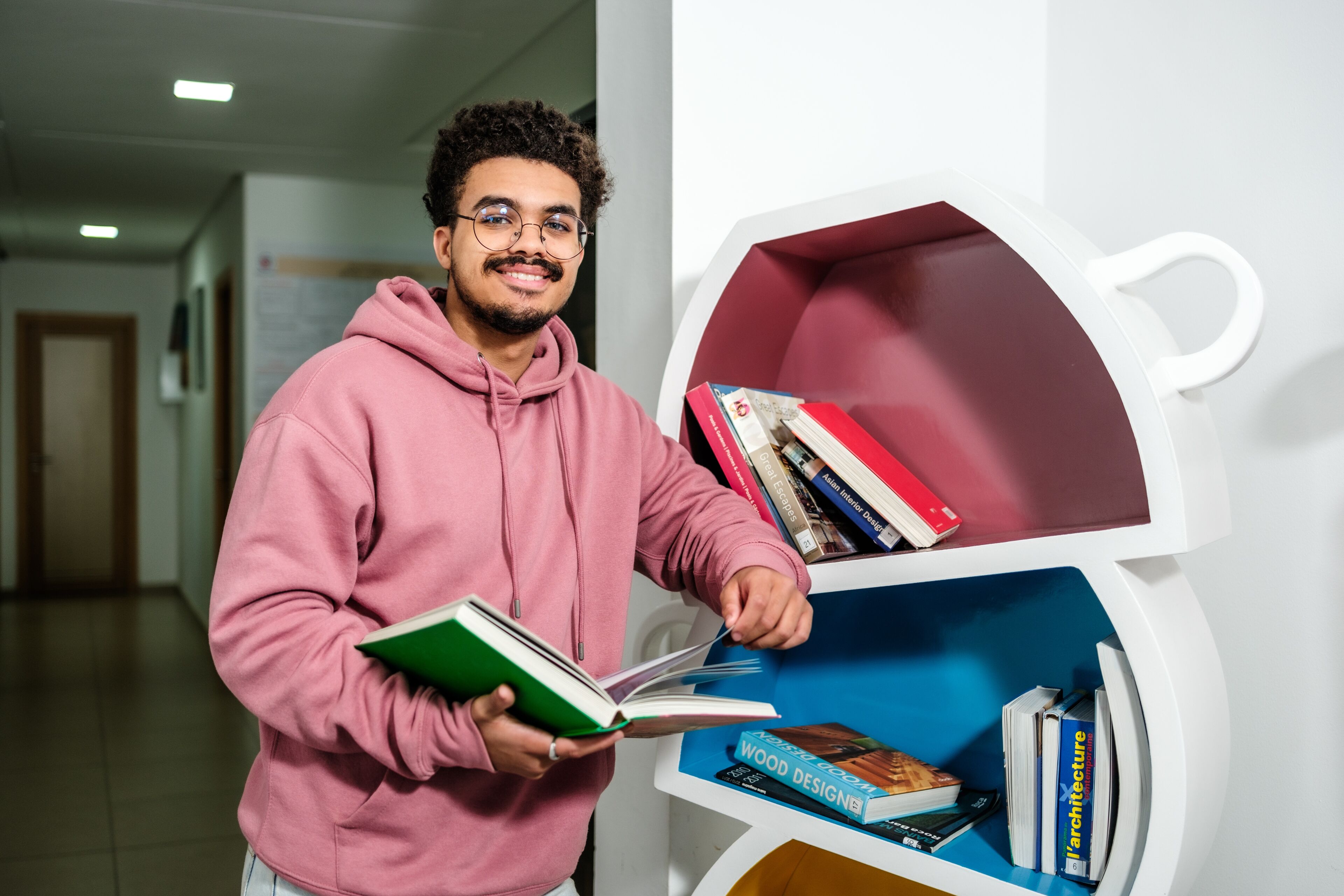 A smiling young man with glasses and a pink hoodie selects a book from a white, cup-shaped bookshelf.