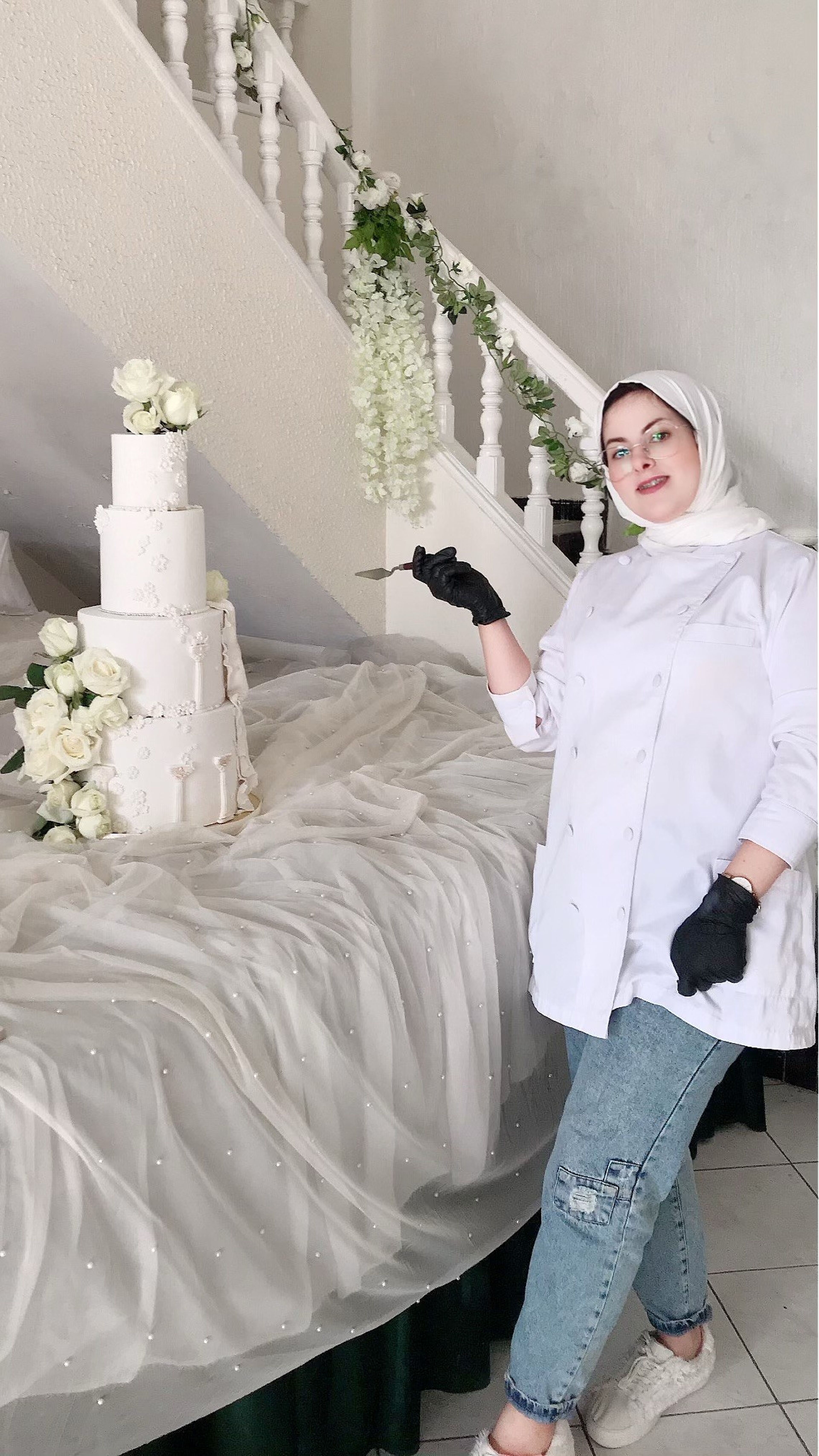 Un chef pâtissier en uniforme blanc et hijab, tenant une spatule, se tient fièrement à côté d'un gâteau de mariage à plusieurs étages orné de roses blanches.