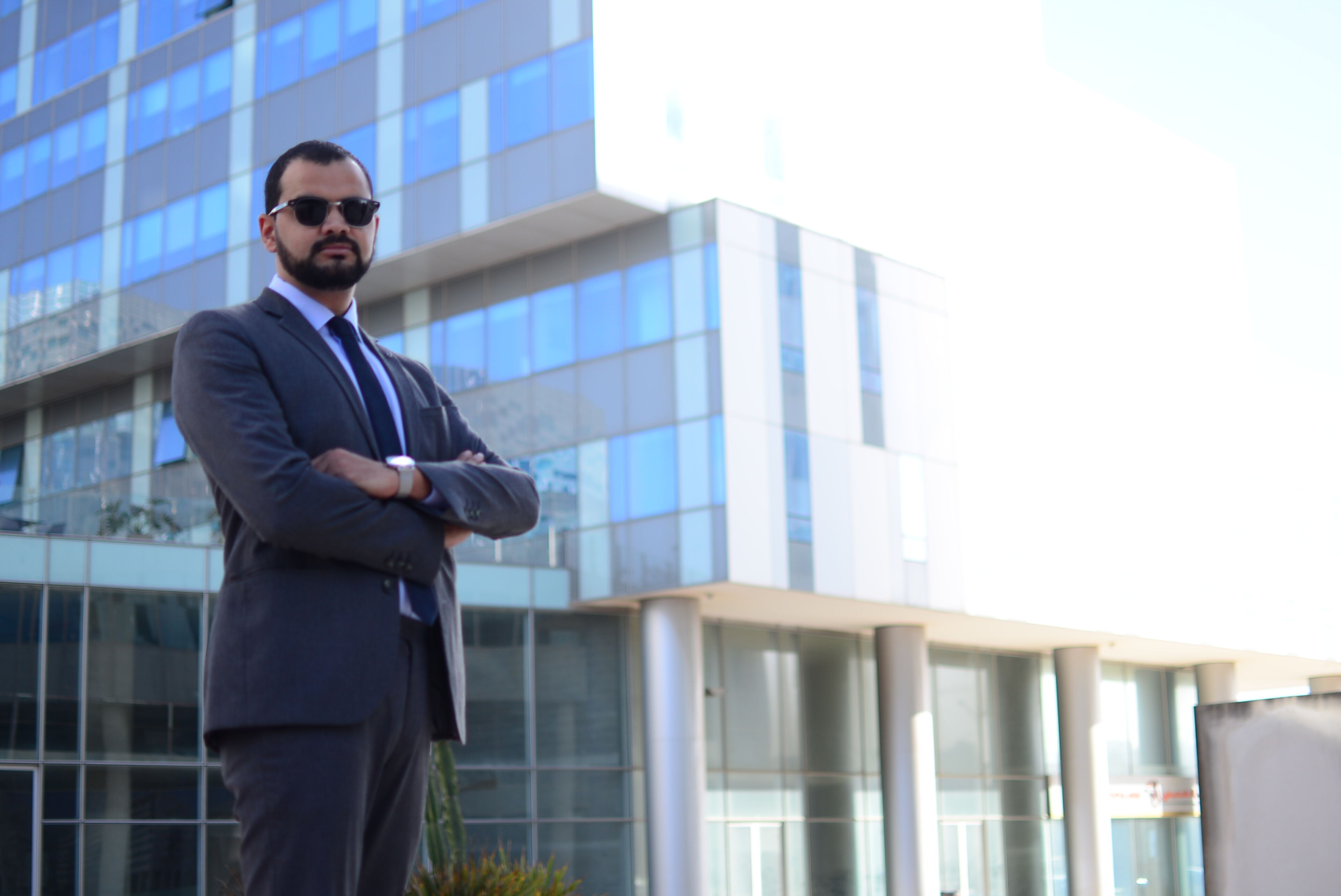 Un homme confiant en costume sombre et lunettes de soleil se tient les bras croisés devant un bâtiment moderne, dégageant une atmosphère professionnelle.
