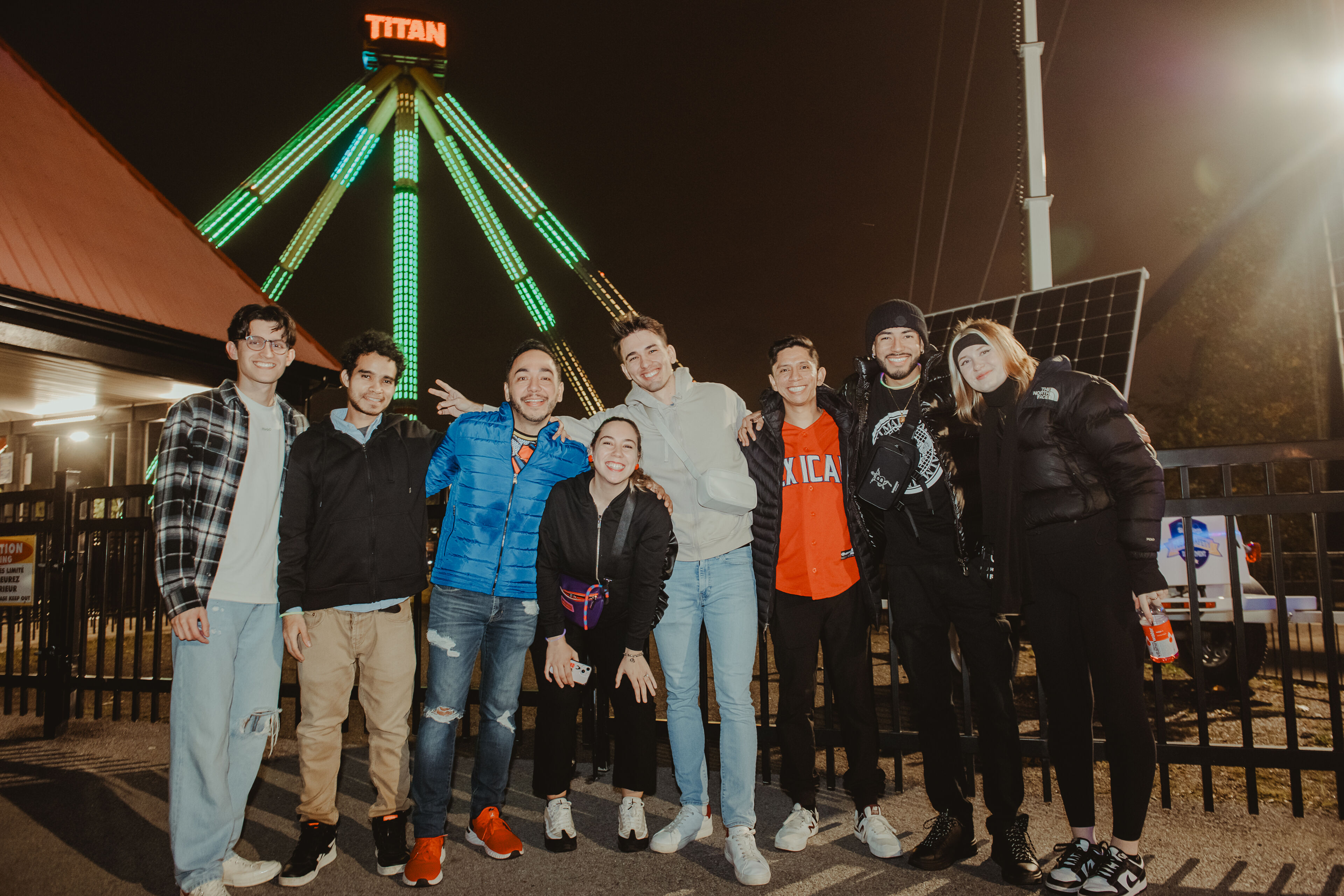 Friends gather for a fun night out, smiling beneath the glowing lights of an amusement park ride.