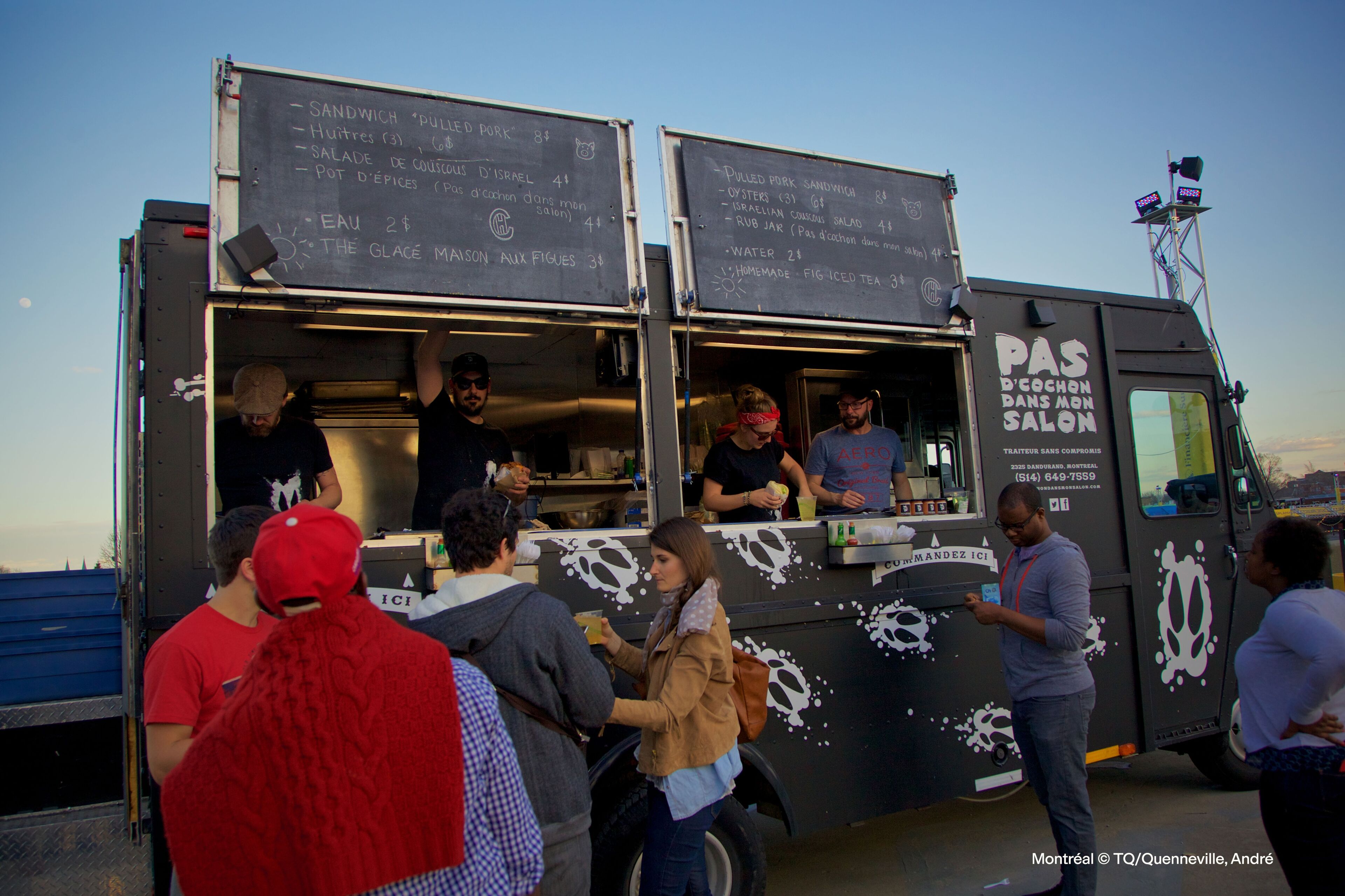 Customers queuing at a food truck serving various dishes and beverages during a sunny evening.