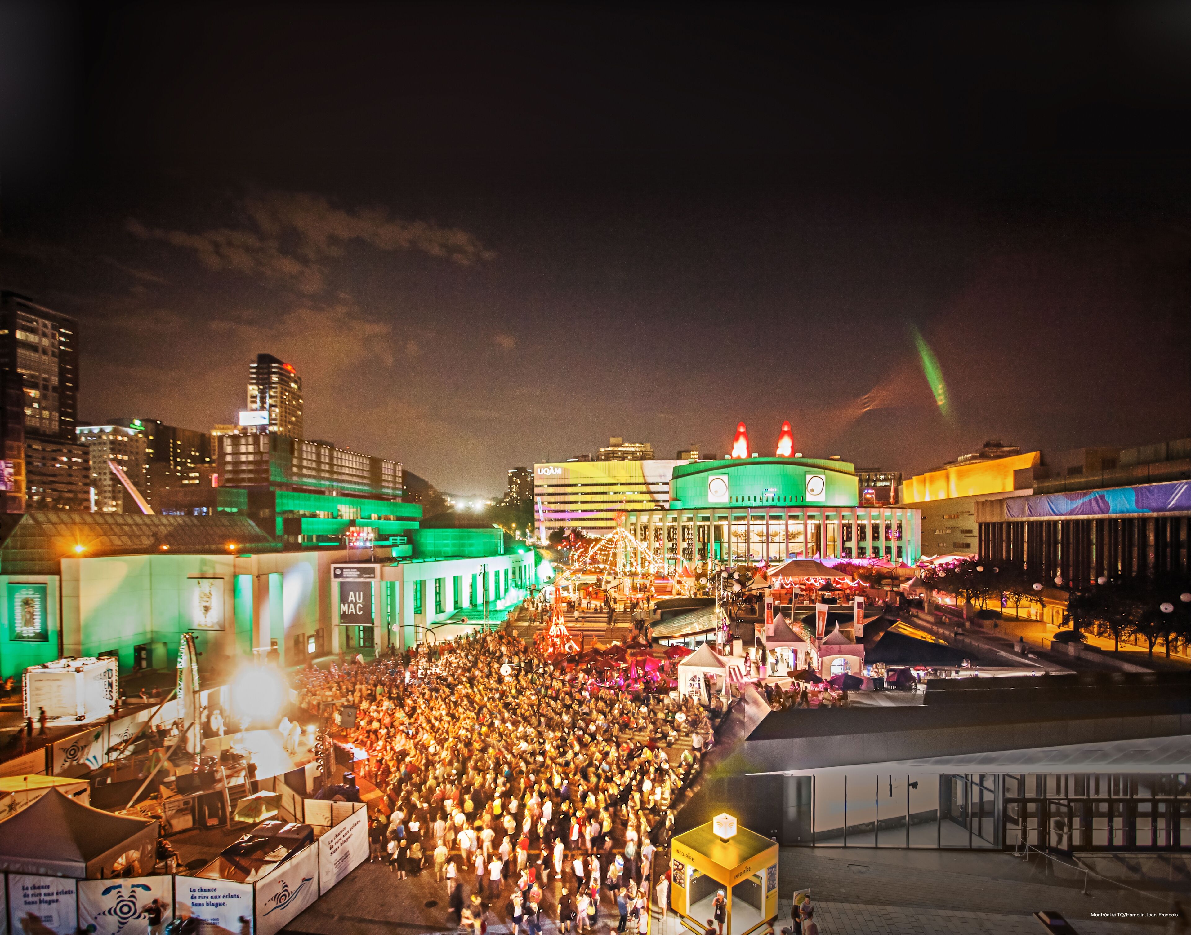 A bustling night festival in the city, with crowds of people gathered around the stage area, vibrant lighting, and city buildings in the background