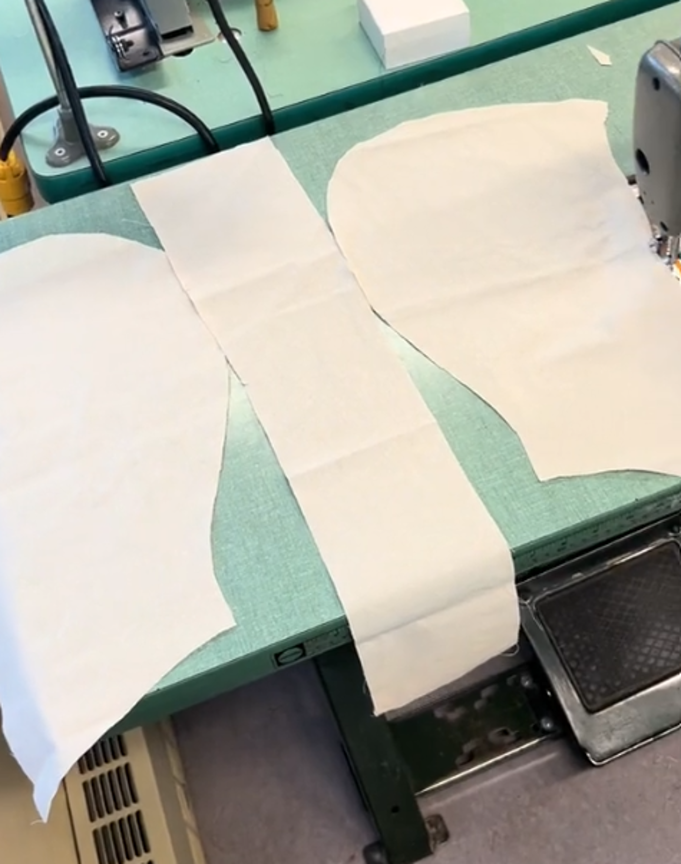 Three pieces of off-white fabric are laid out on a teal sewing table next to an industrial sewing machine.