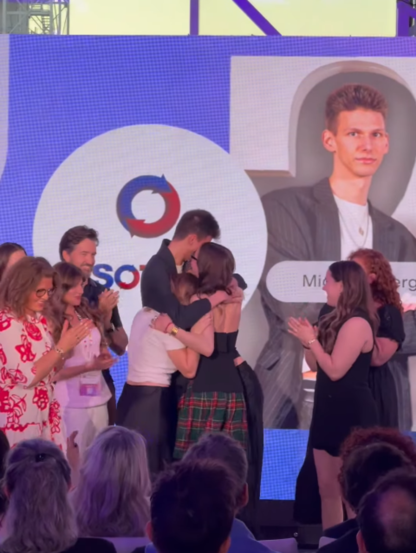 A group of people, mostly women, are embracing on a stage, with others watching and applauding.