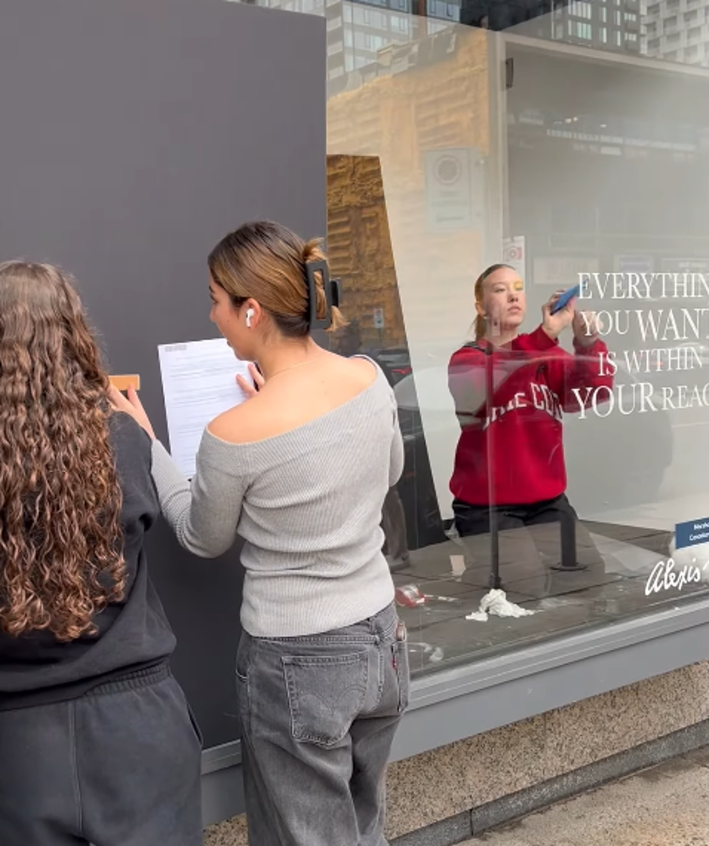 Two women stand in front of a dark gray wall, one holding a piece of paper while the other appears to be taking a picture of a store window displaying the words "Everything you want is within your reach."