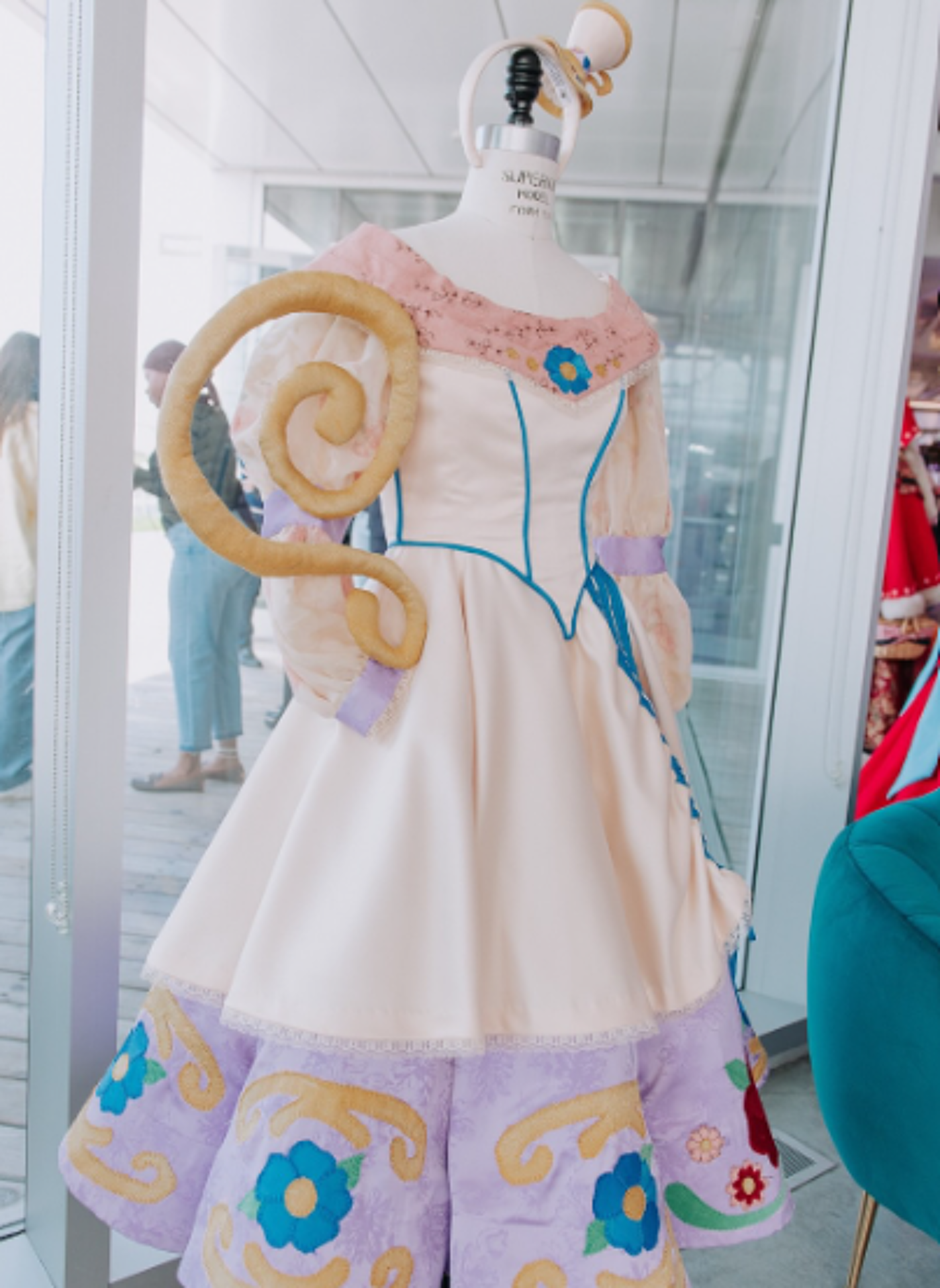 A cream-colored dress with lavender and teal accents, adorned with embroidered flowers and a unique, swirling, breadstick-like accessory, is displayed on a mannequin in front of a glass door.