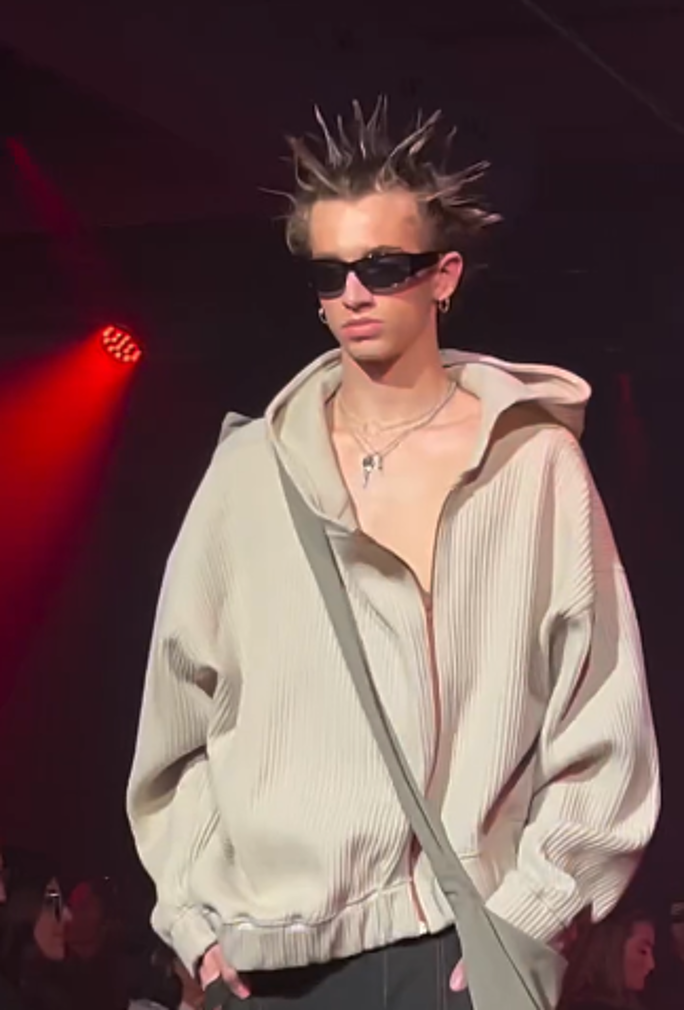 A young man with spiky hair, sunglasses, and a beige hooded jacket walks a runway.