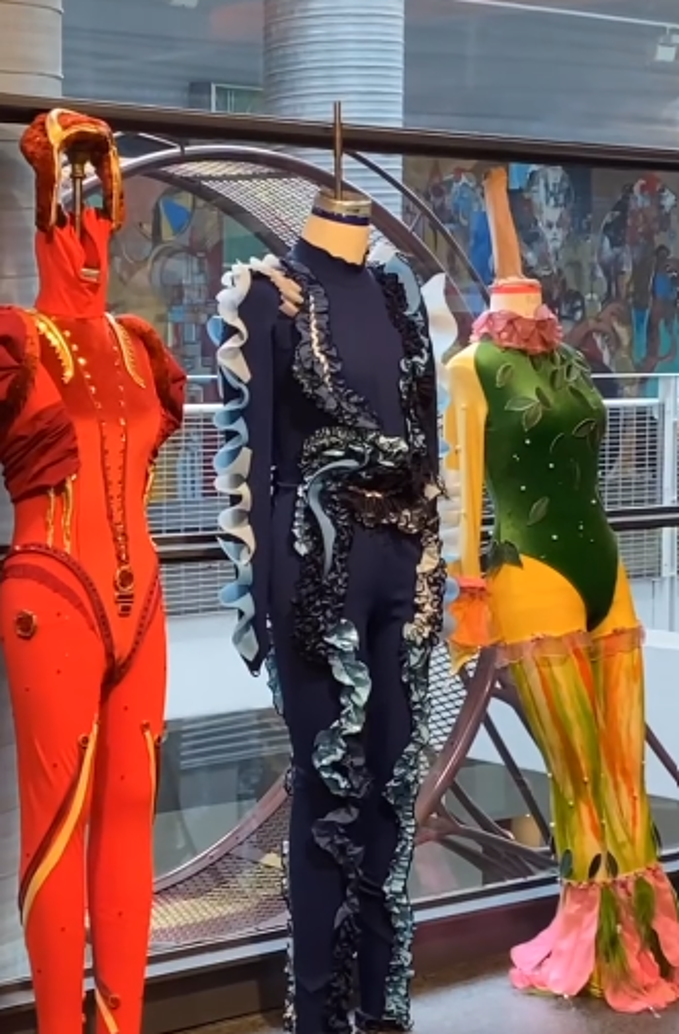 Three mannequins display vibrant, uniquely designed costumes, showcasing detailed craftsmanship and bold color palettes against a backdrop of a modern building's glass facade and mural.