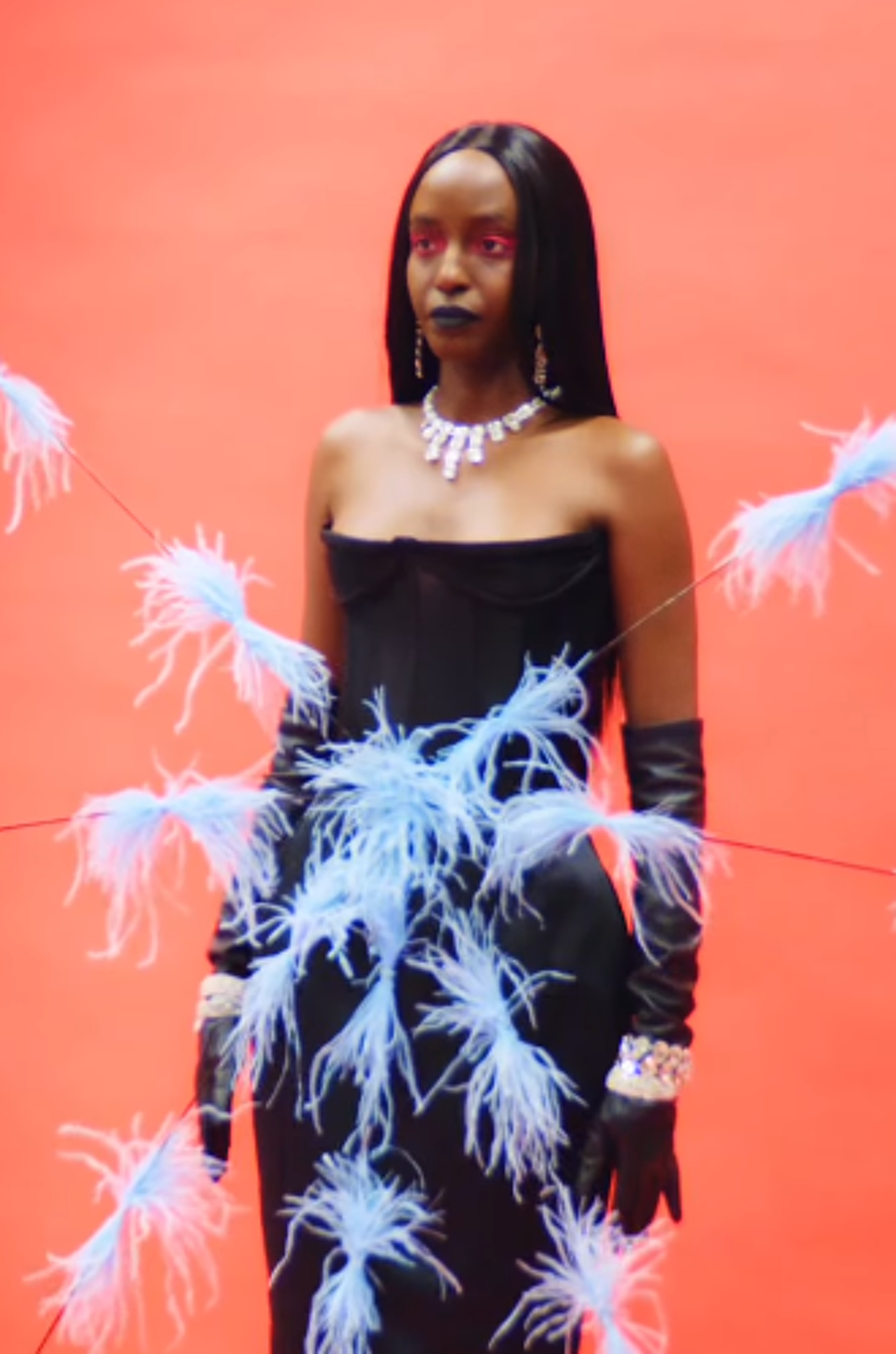 A Black woman with long black hair and pink eyeshadow wears a black strapless dress adorned with light-blue feathers and long black gloves against a coral background.